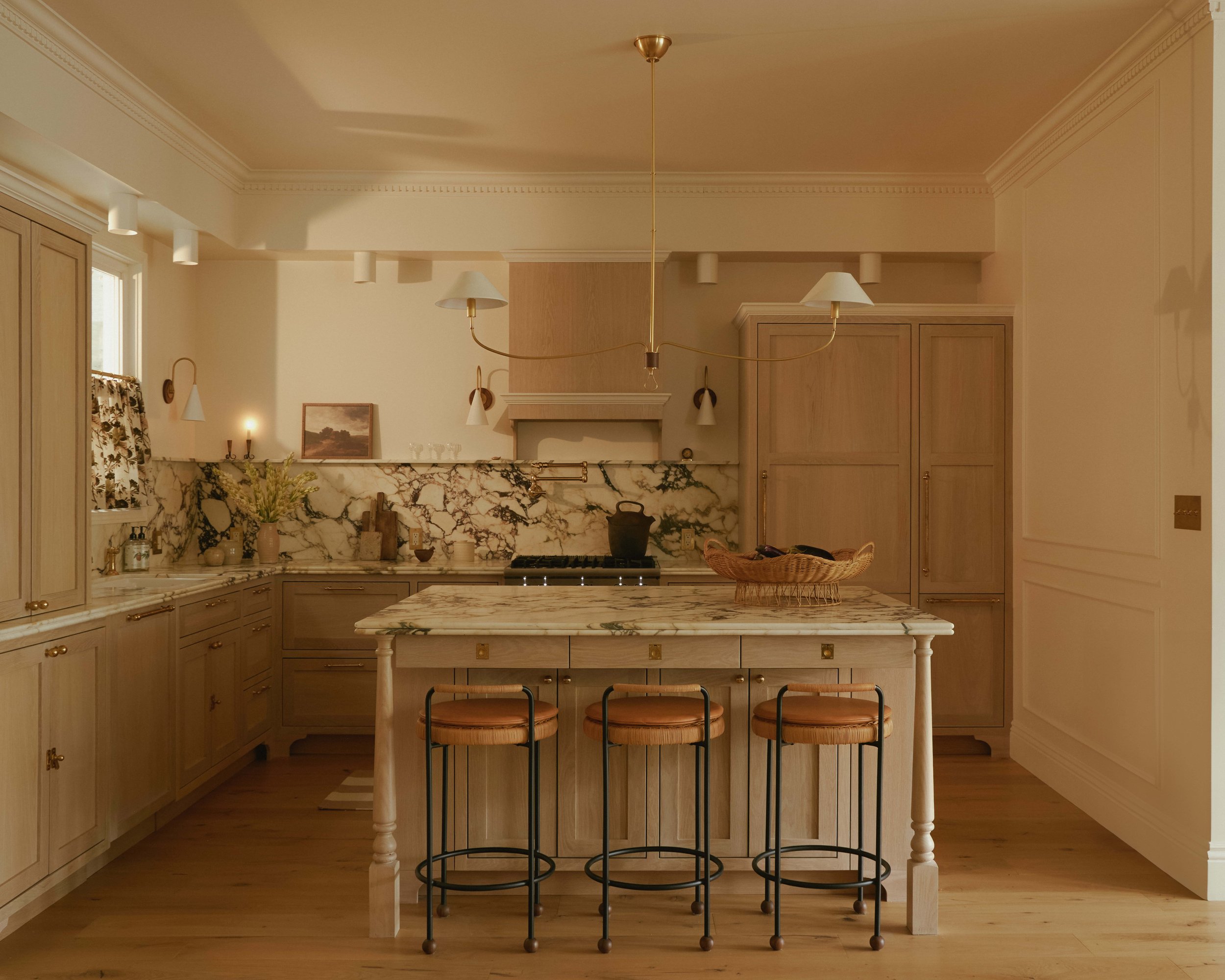 A warm, marble-clad kitchen island with brass pendant lighting and rattan counter stools
