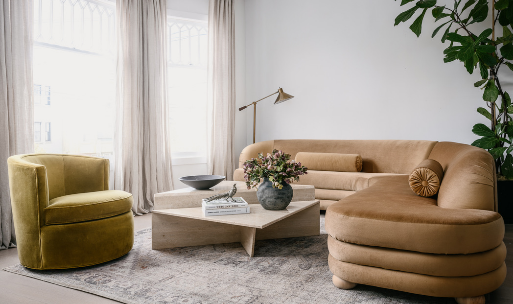 Living room with a curved velvet sectional sofa, mustard swivel armchair, square travertine coffee table, and brass floor lamp, gothic-arched windows, styled with fresh flowers and a bronze figurine, San Francisco gut renovation by Spearman Spaces