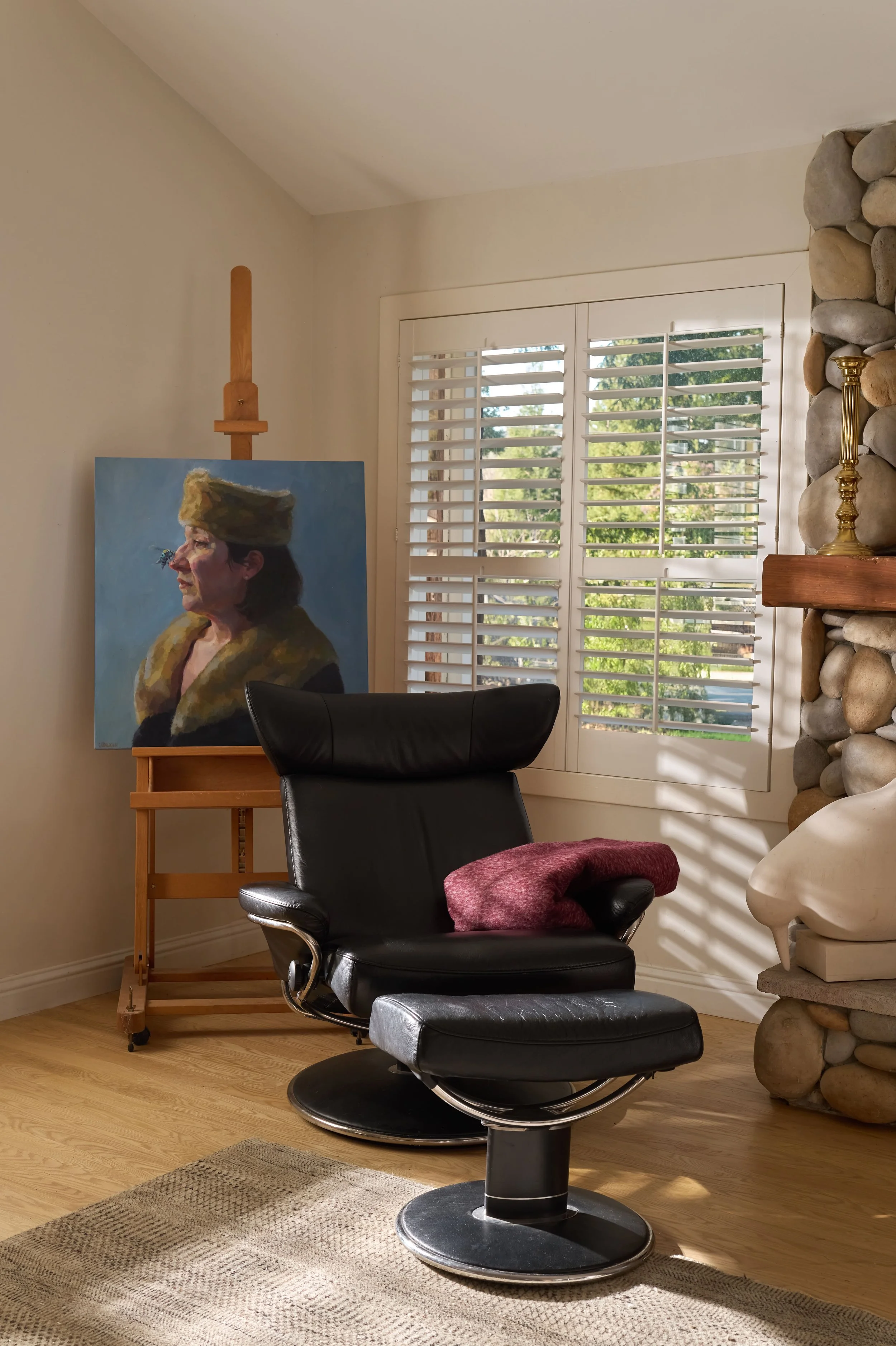 Black leather recliner with ottoman and a maroon throw beside an oil portrait on a wood easel, with a river-rock stone fireplace edge, brass candlestick on a wood mantel, and a sculptural dove object, Alamo Farmhouse whole-home remodel (Jackson Way, 