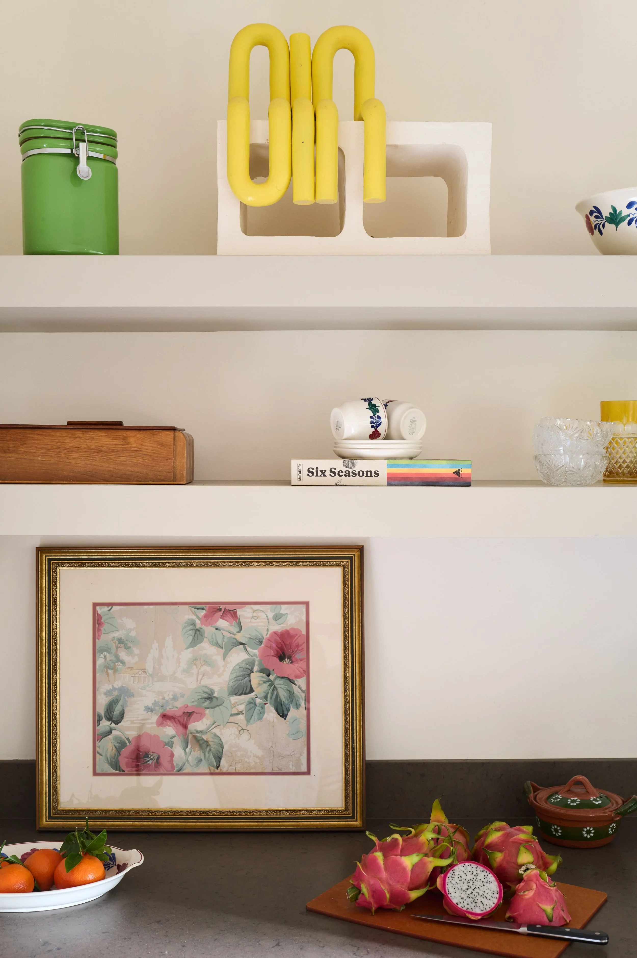 Built-in shelving with books and bowls above a kitchen counter styled with framed floral art and dragon fruit being sliced on a wood cutting board, Alamo Farmhouse (Jackson Way) complete interior remodel by Spearman Spaces