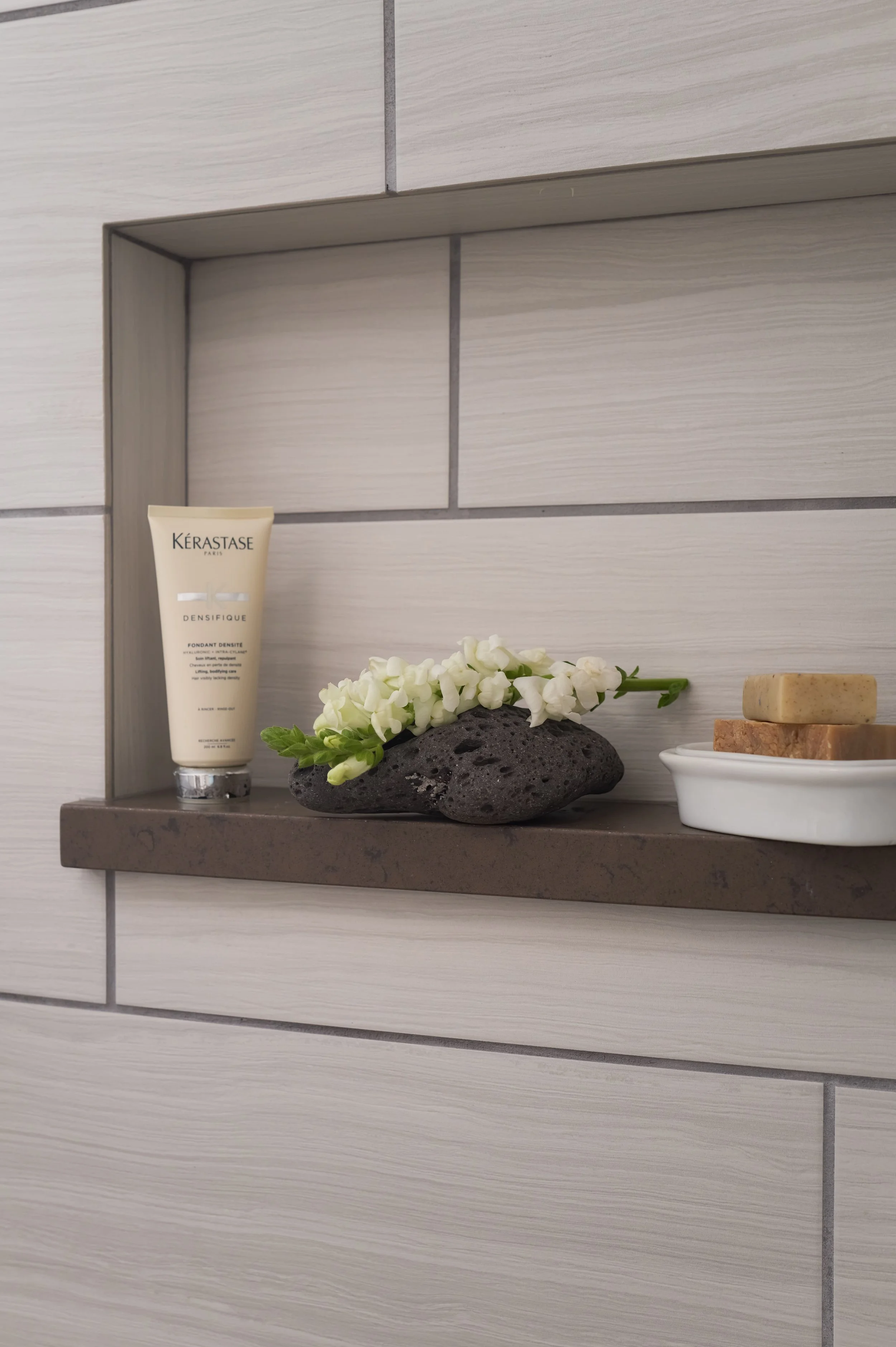 Shower niche shelf styled with Kerastase hair product, a lava stone vessel with white snapdragon blooms, and a stack of soap bars on a white dish, set into gray wood-look tile, Alamo Farmhouse (Jackson Way) pool house by full-service interior design 