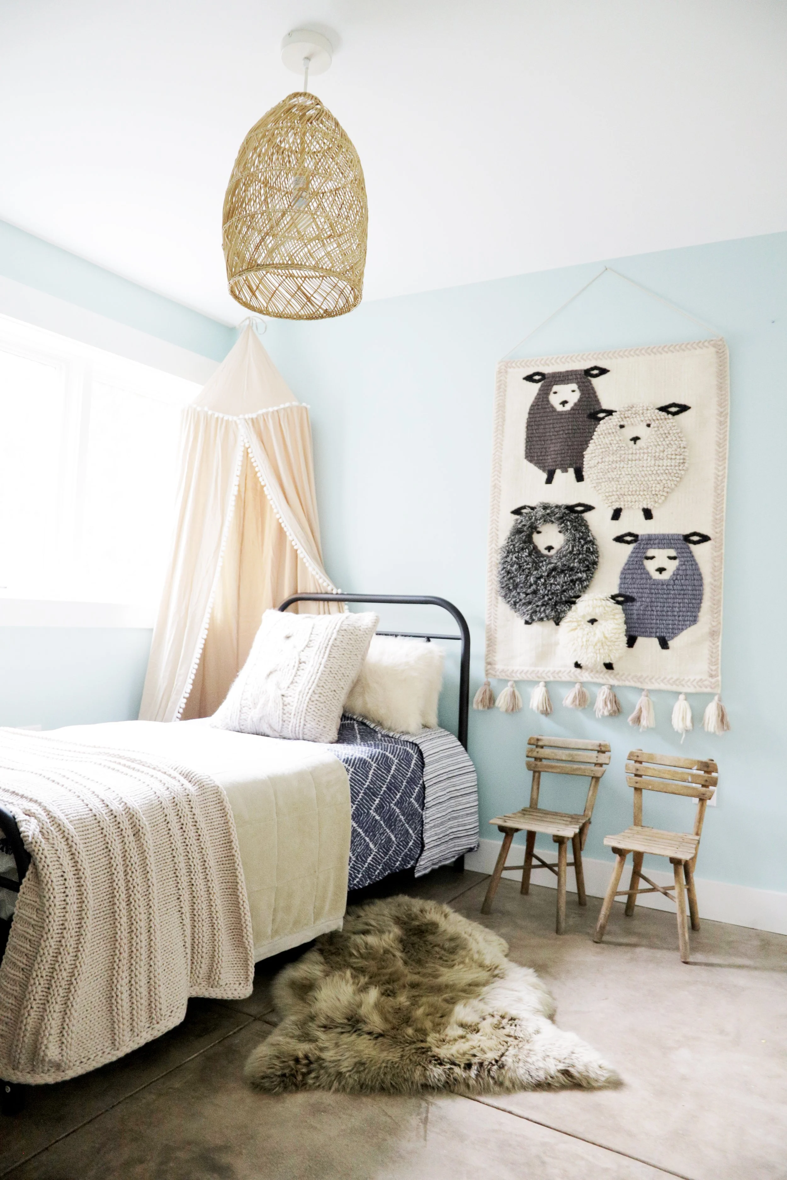 Kids bedroom with pale blue walls, a black iron daybed layered in cream knit and striped bedding, a woven wall hanging of sheep with tassel trim, a rattan pendant light, a pink canopy drape, a sheepskin rug, and small vintage wood chairs, Oakland Bun