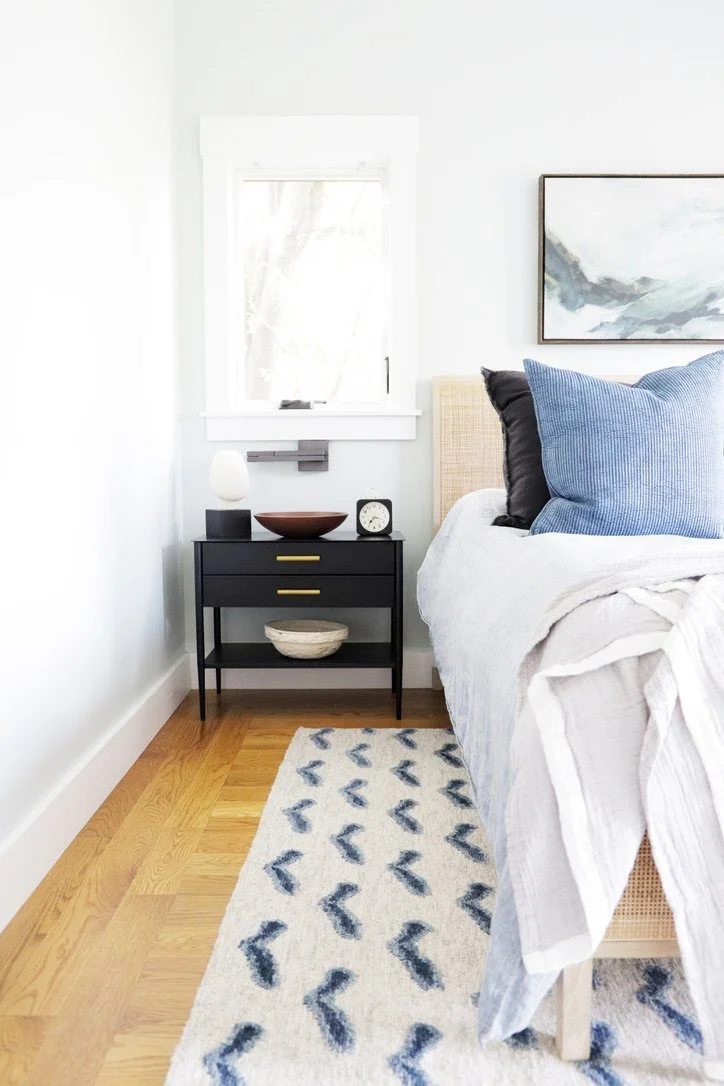Bedroom nightstand detail with a black wood nightstand with brass pulls, a white ceramic table lamp, a walnut bowl, and a cane headboard dressed in blue and charcoal pillows, Oakland Bungalow (61st Street) full-service remodel by Spearman Spaces