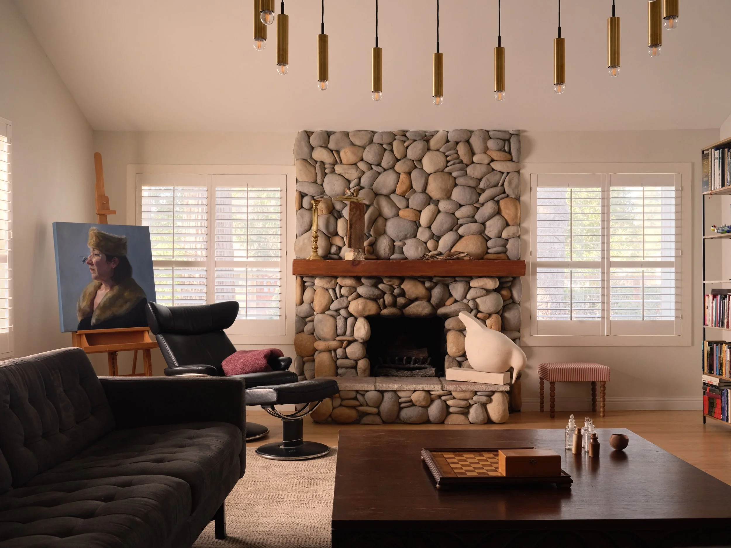Living room with river-rock stone fireplace, wood mantel, black leather recliners, and an oil portrait on an easel, Alamo Farmhouse (Jackson Way) full-service remodel by Spearman Spaces