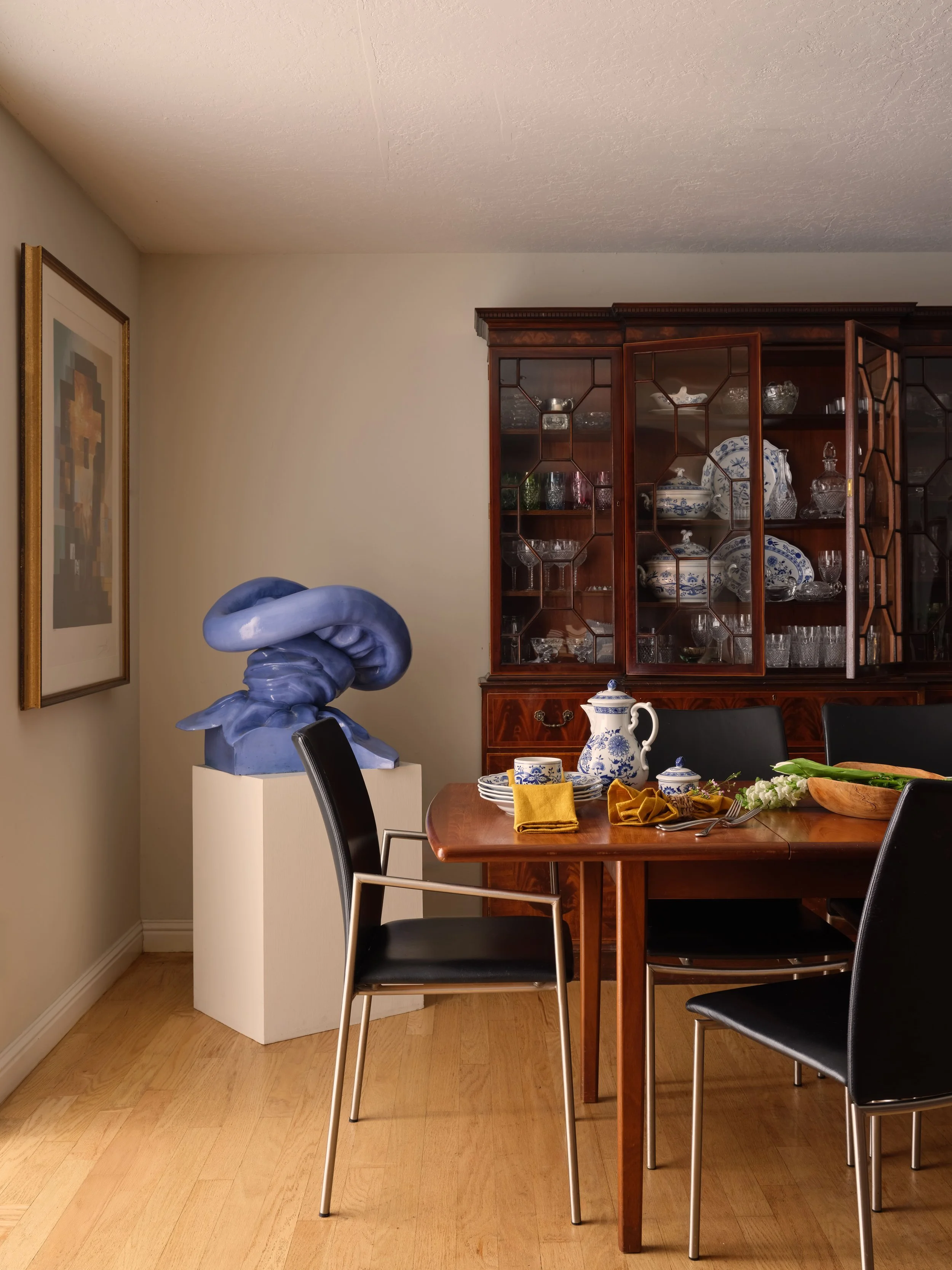 Dining room with a sculptural blue ceramic knot form on a white pedestal, an antique mahogany china cabinet displaying blue and white porcelain, a wood dining table set with mustard linen napkins and fresh snap peas, and black leather dining chairs, 