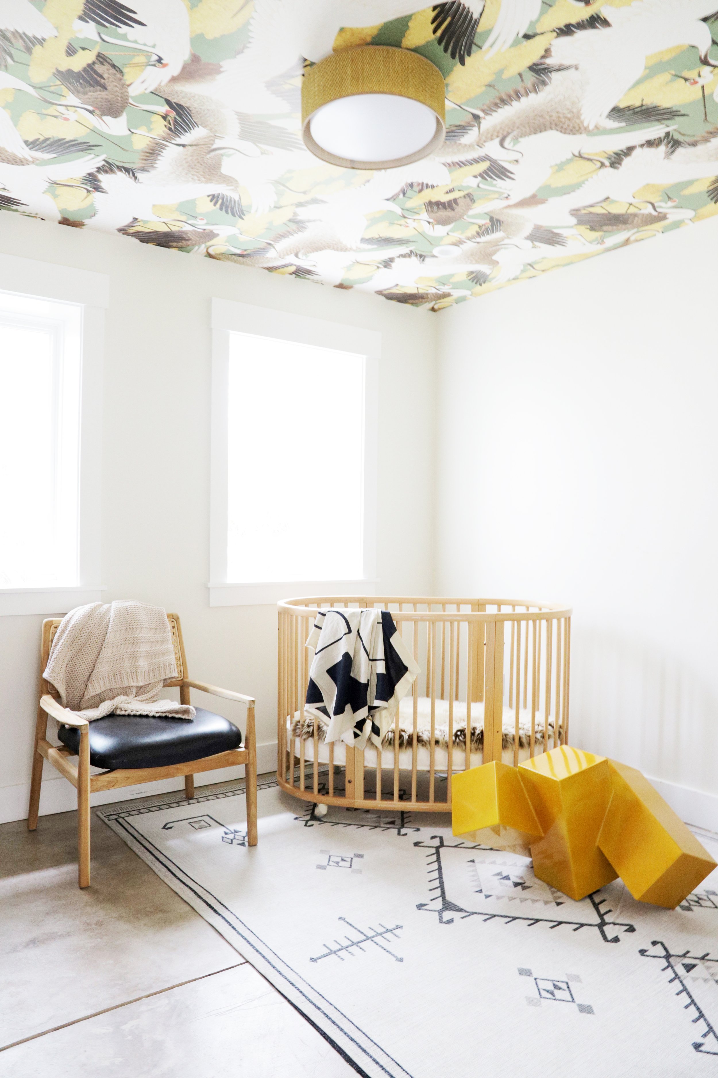 Nursery with crane and cloud motif wallpaper on the ceiling, a gold flush-mount light, round wood spindle crib, cane-and-leather accent chair, and a sculptural geometric yellow object on a black and white patterned rug, Oakland Bungalow (61st Street)