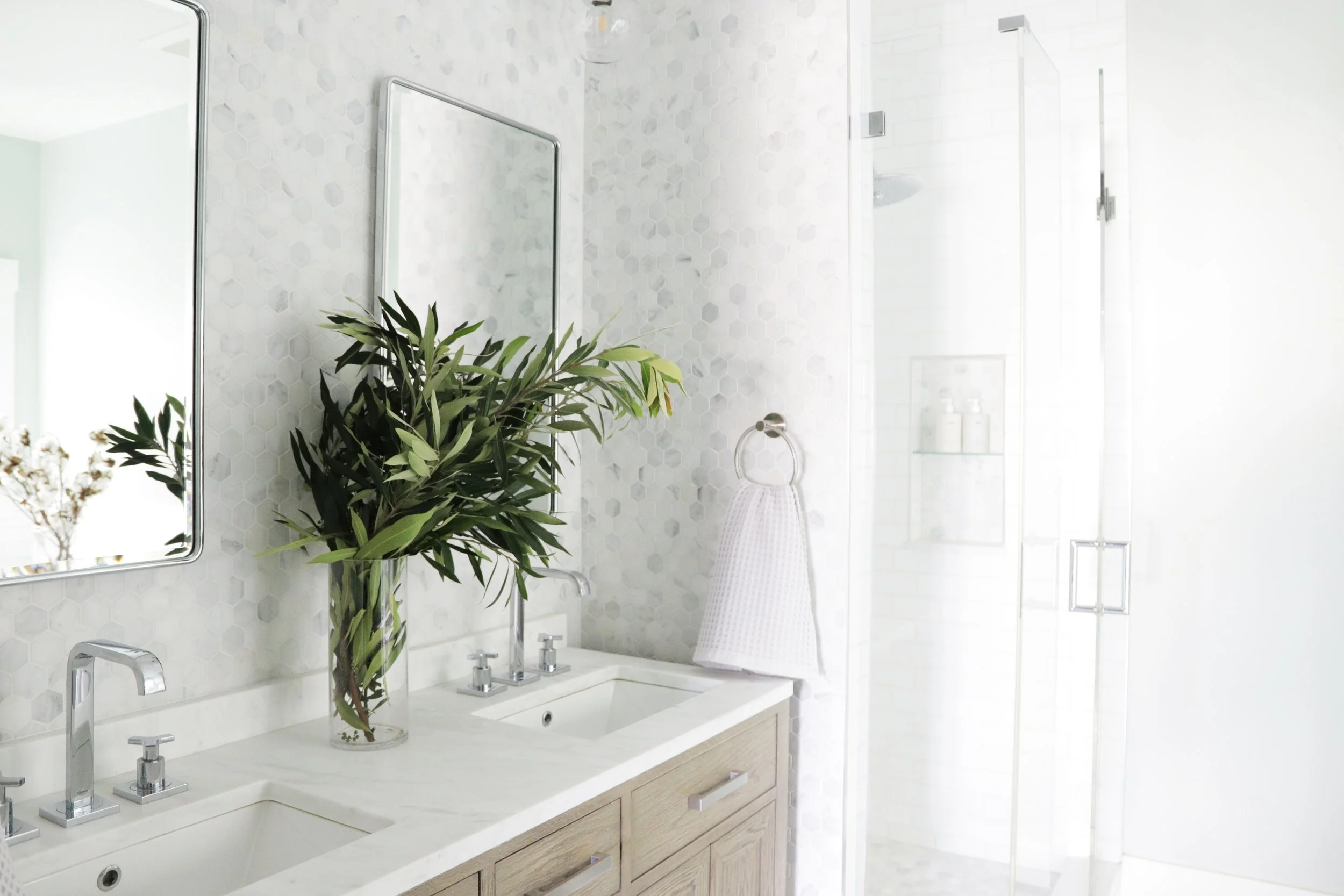 Primary bathroom with a white oak double vanity, marble countertop, gray hexagon marble tile walls, a glass-enclosed shower with a built-in niche, and fresh olive branches in a glass vase full-service interior remodel by Spearman Spaces