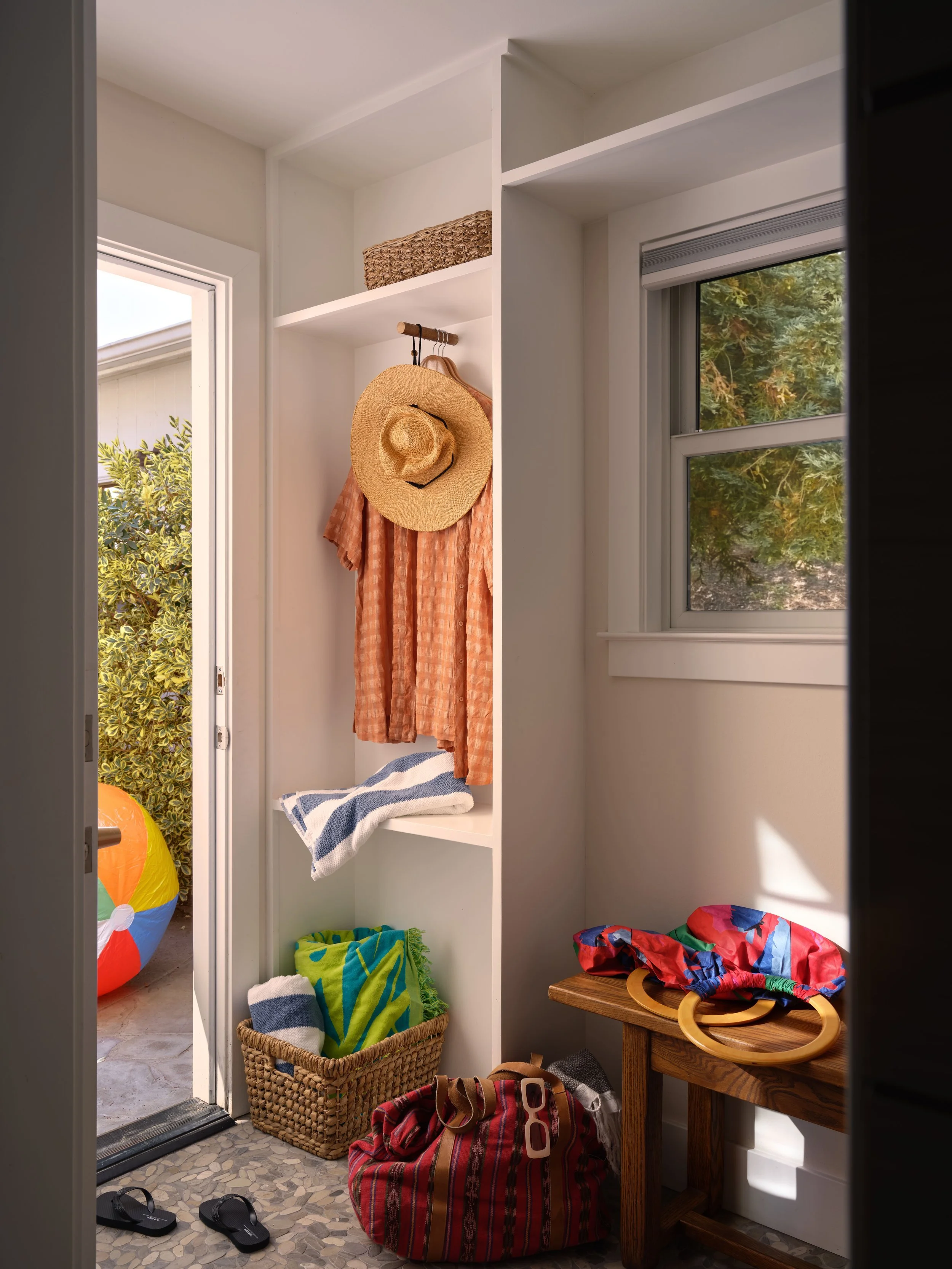 Pool house entryway with a built-in coat nook holding a straw sun hat, a coral robe, a striped towel, and a woven storage basket, a wood bench styled with colorful tote bags, and an open door leading outside to a beach ball and garden hedge, Alamo Fa