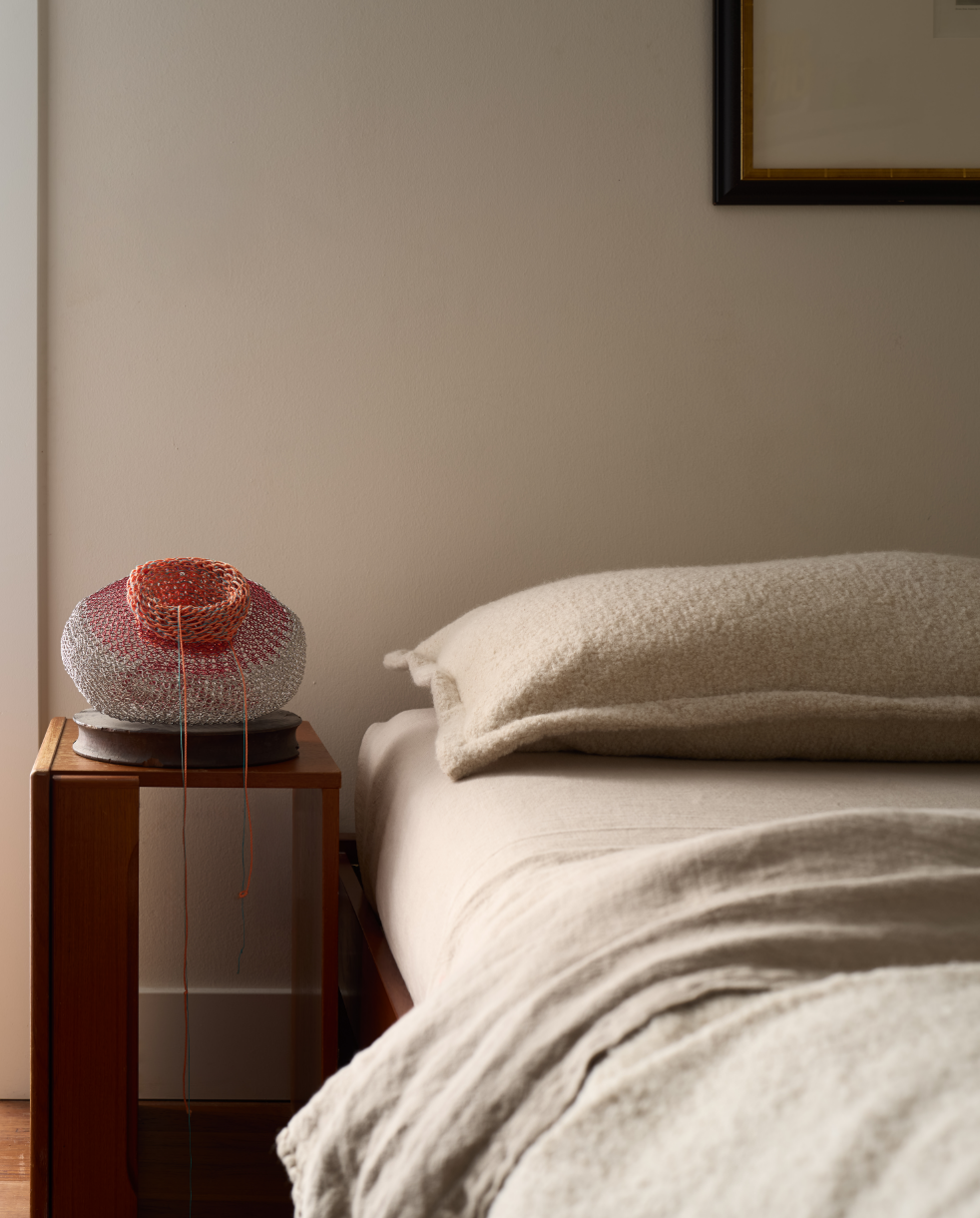 Bedroom nightstand with a red and cream woven table lamp on a mid-century wood nightstand, beside a bed layered in boucle and linen bedding, Alamo Farmhouse remodel (Jackson Way, Alamo) by full-service interior design studio Spearman Spaces