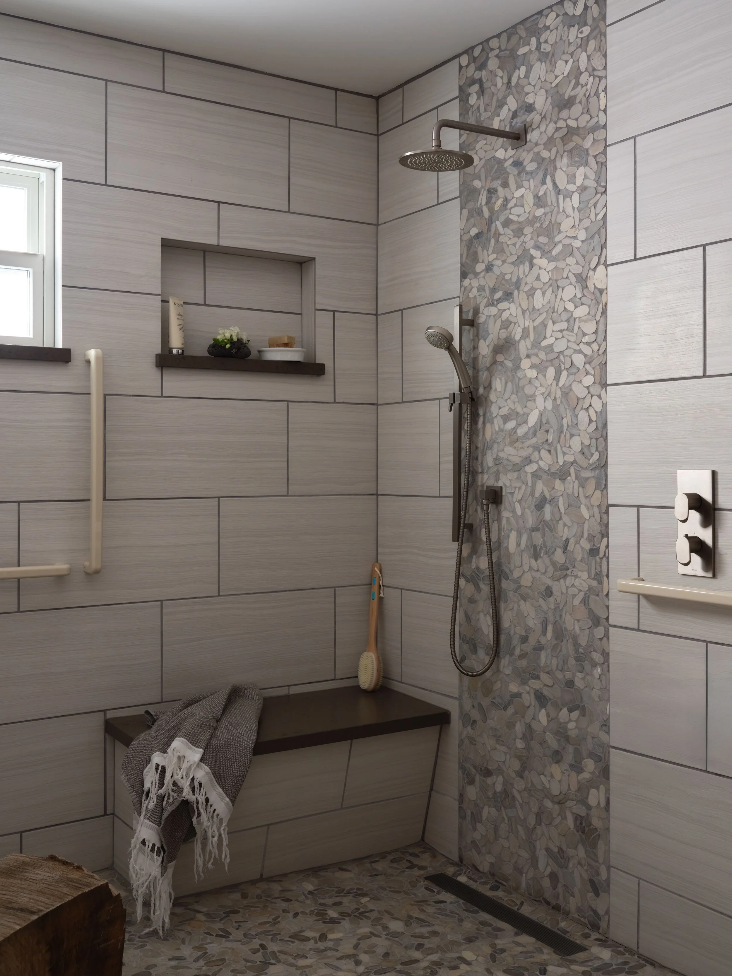 Zero-threshold roll-in shower with oil-rubbed bronze grab bars, a rainfall shower head, a handheld shower wand, a river-stone pebble accent tile strip, and a built-in tile bench, aging-in-place design in the Alamo Farmhouse (Jackson Way) pool house b