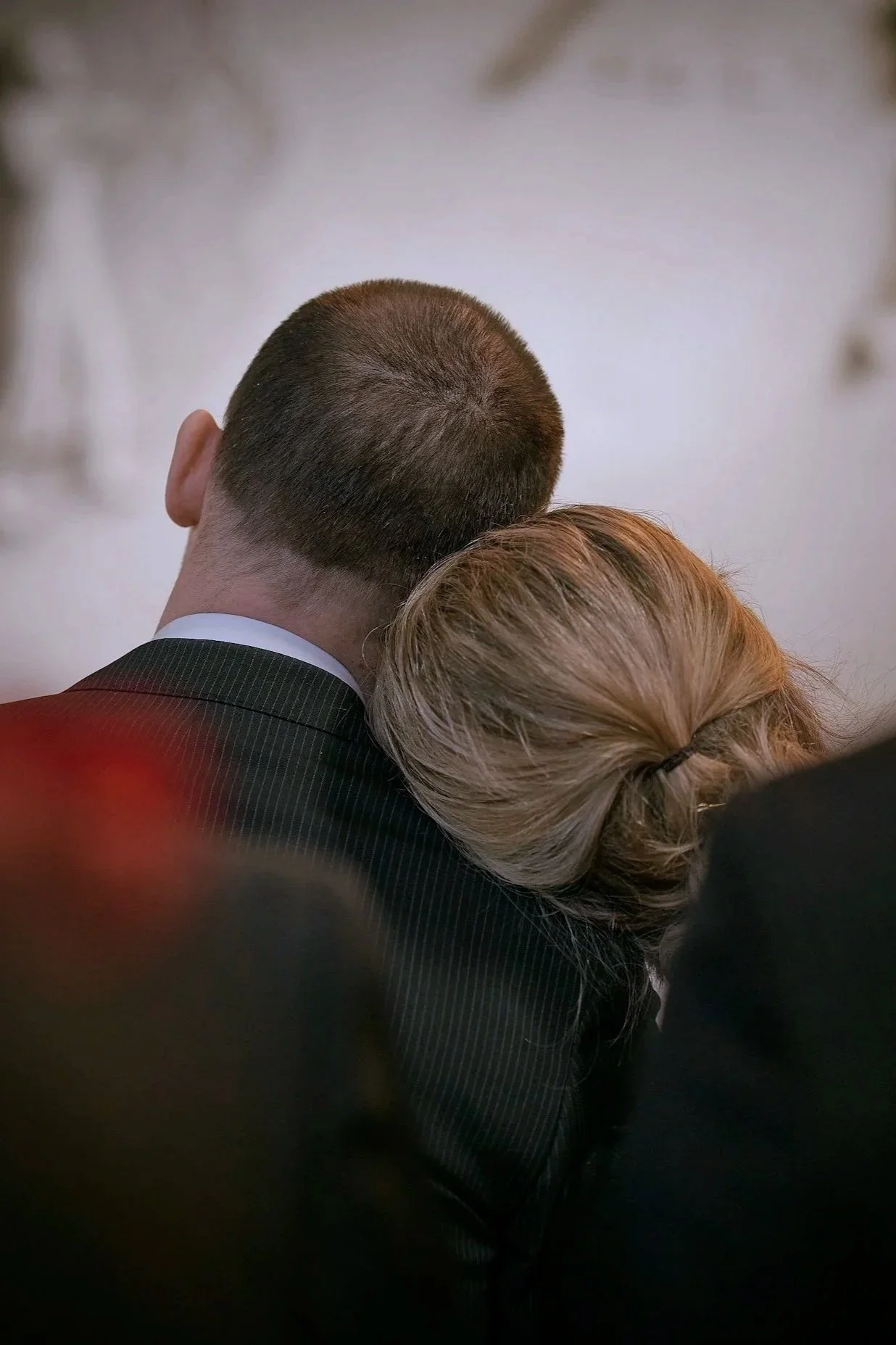 A man in a suit with short dark hair and a woman with light brown hair and a hair clip, resting their heads together