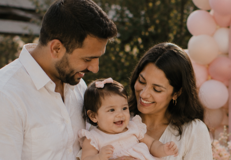 A happy family of three with a father, mother, and baby girl smiling and looking at each other outdoors, with pink and white balloons in the background at a celebration or outdoor event.