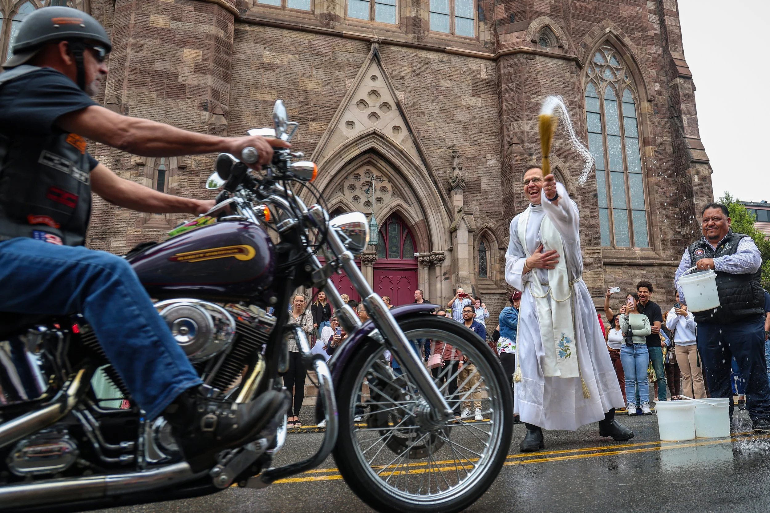 PATERSON, N.J. | May 2, 2025 — Rev. Msgr. Geno R. Sylva blesses passing motorcyclists with holy water as riders cruise past the Cathedral of St. John the Baptist, as churchgoers and neighborhood residents line the sidewalks, snapping photos and cheer