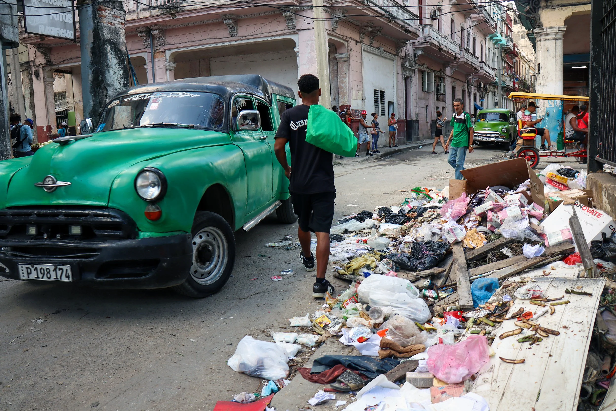 HAVANA, CUBA — March 9, 2026: A man squeezes through a narrow gap between a pile of garbage and a classic American car on a corner in Central Havana (Centro Habana). Recent layoffs of government sanitation workers have contributed to mounting trash i