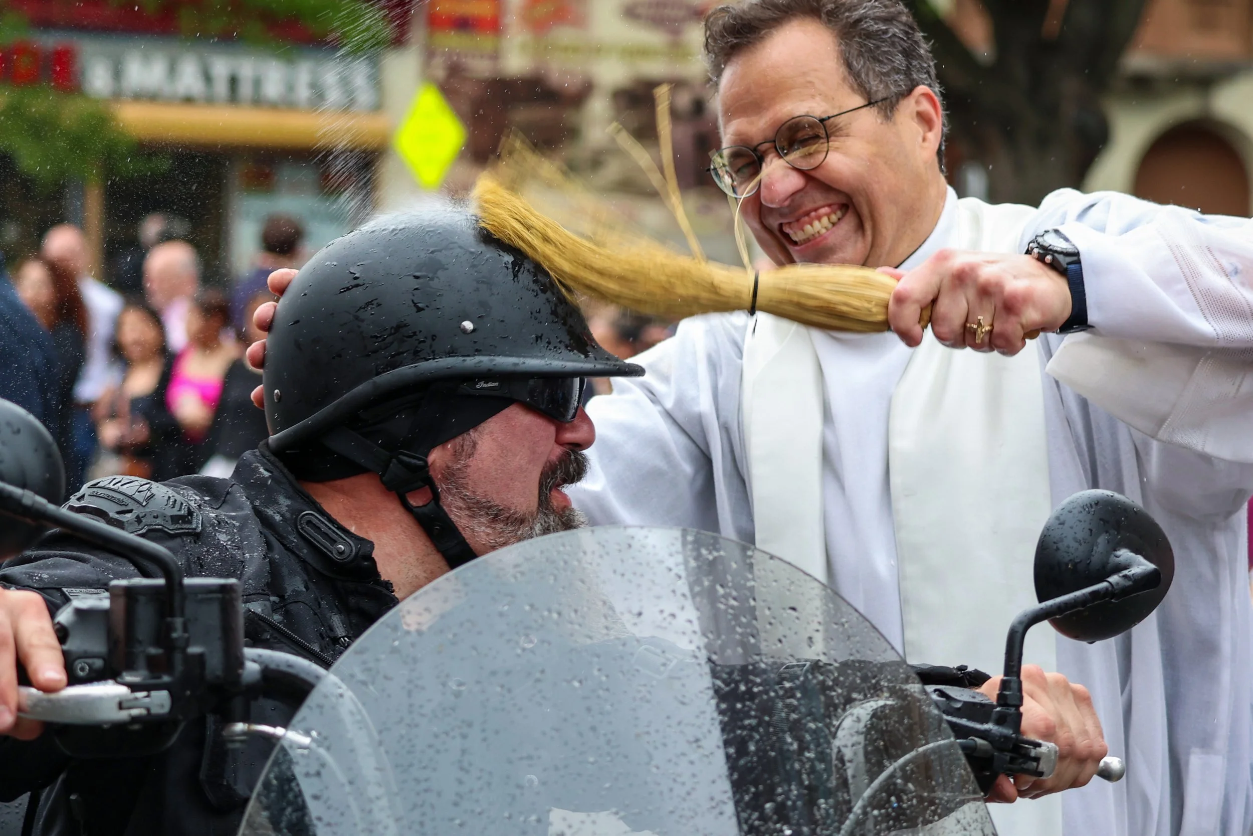 PATERSON, N.J. | May 2, 2025 — Rev. Msgr. Geno R. Sylva splashes holy water on a biker’s helmet as riders cruise past the Cathedral of St. John the Baptist, while churchgoers and neighborhood residents line the sidewalks, snapping photos and cheering