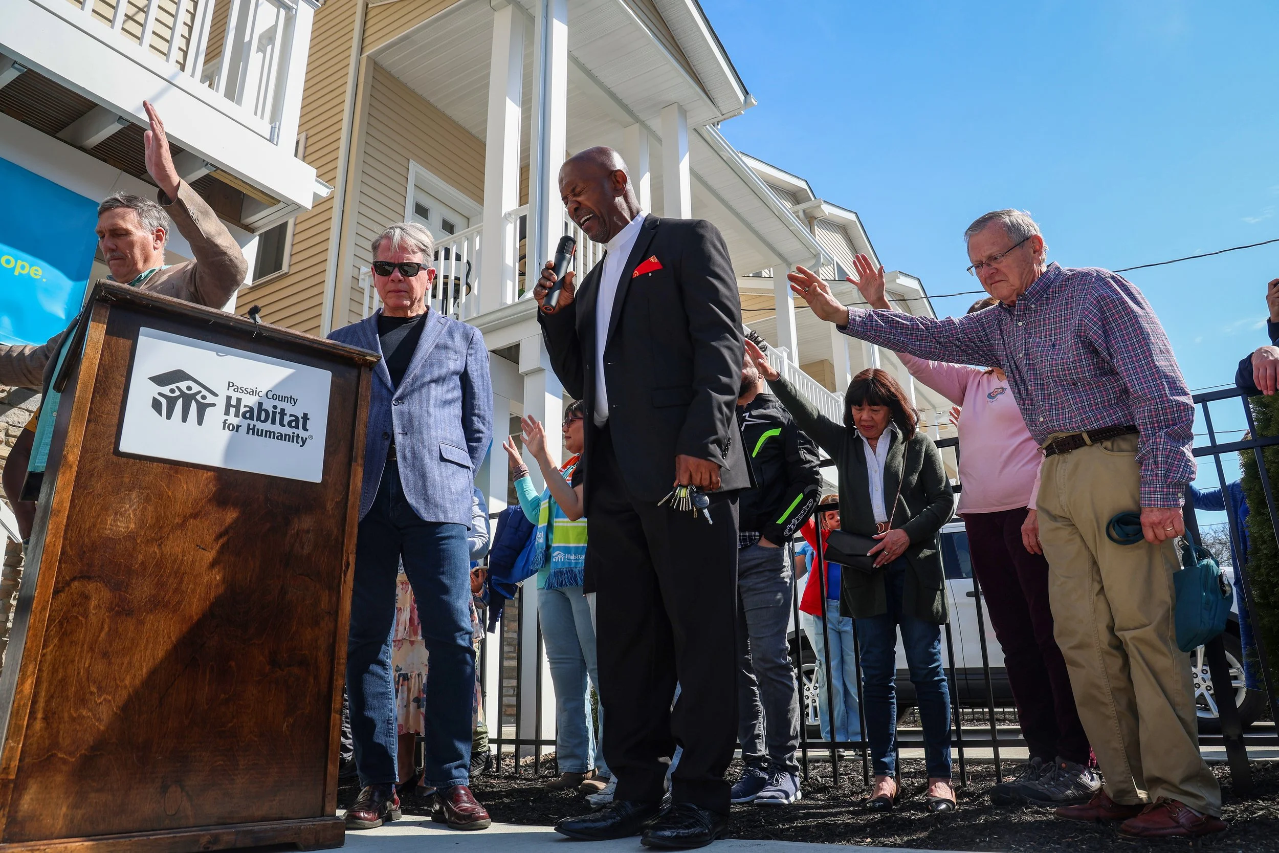 PATERSON, N.J. | March 29, 2025 — Rev. Brian Collins offers a blessing as the crowd raises their arms in unison in gratitude, marking the moment when eight Paterson families achieved homeownership through Passaic County Habitat for Humanity. The newl