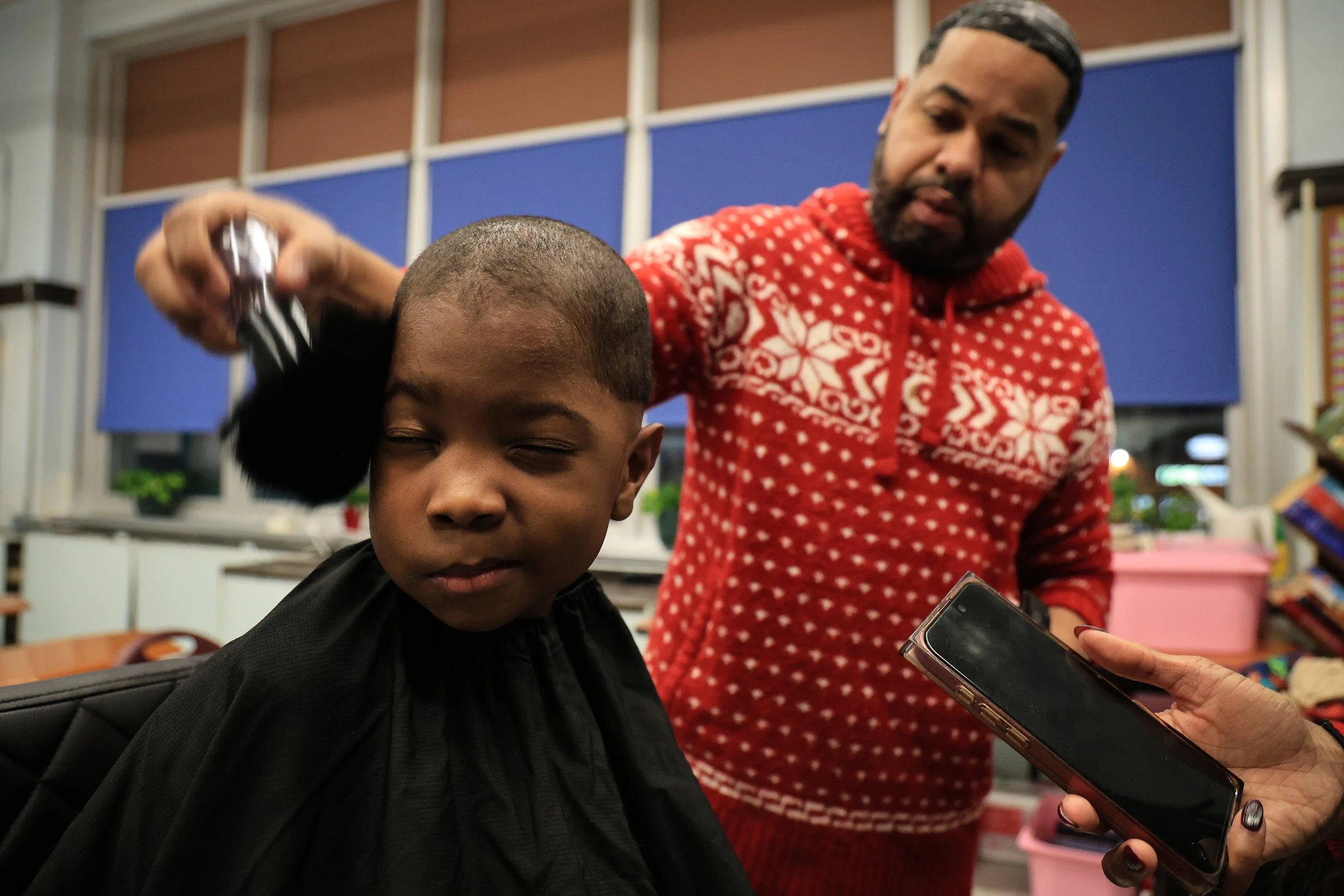 PATERSON, N.J. | 12/17/25: Four-year-old Zaire (last name not given) receives a face brush-off after getting a haircut from barber Jonathan Vicioso of House of Experts Barbershop in Paterson, during the annual holiday dinner at Eastside High School, 
