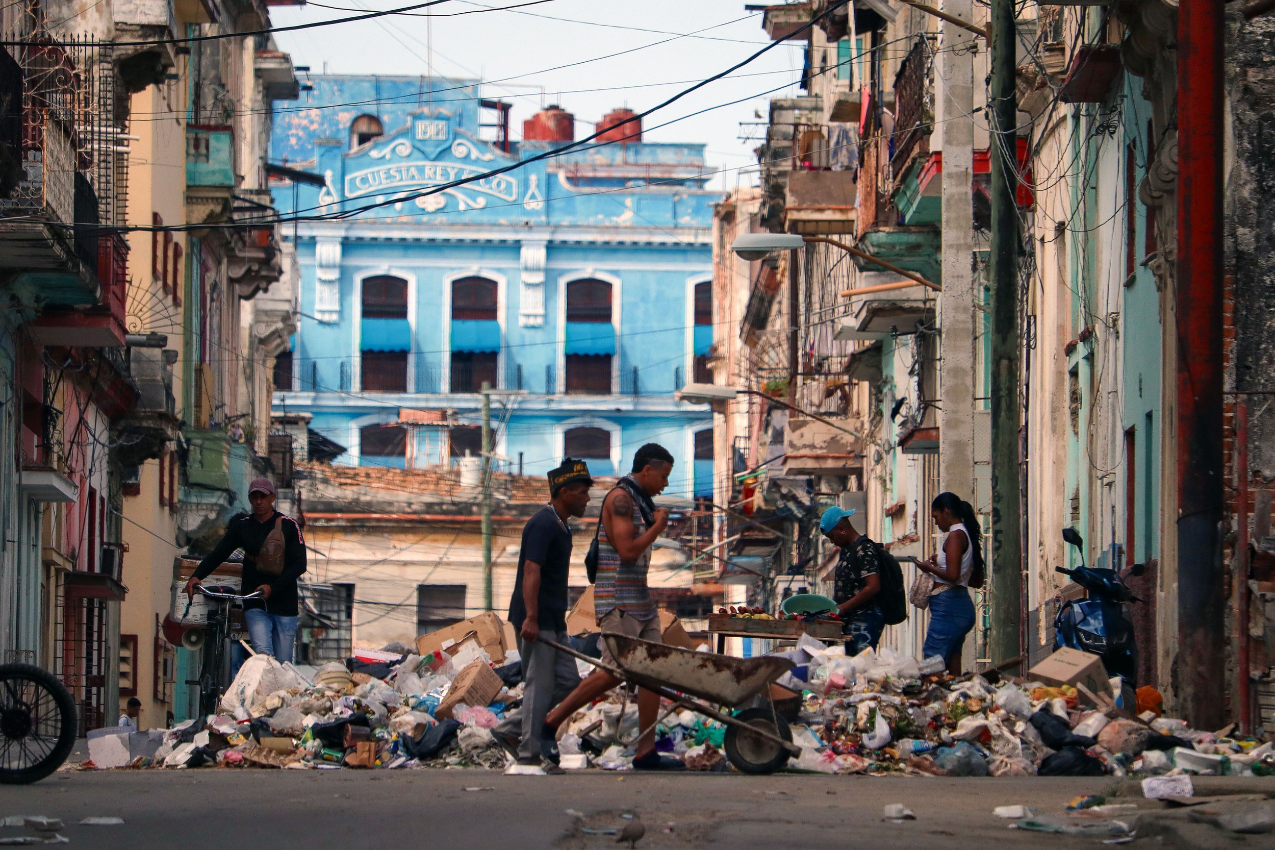 HAVANA, CUBA — March 9, 2026: Two men push a wheelbarrow through Old Havana (Habana Vieja) as others go about their daily lives, navigating streets blocked by piles of trash that create unsafe, unsanitary conditions and disrupt traffic. Recent layoff