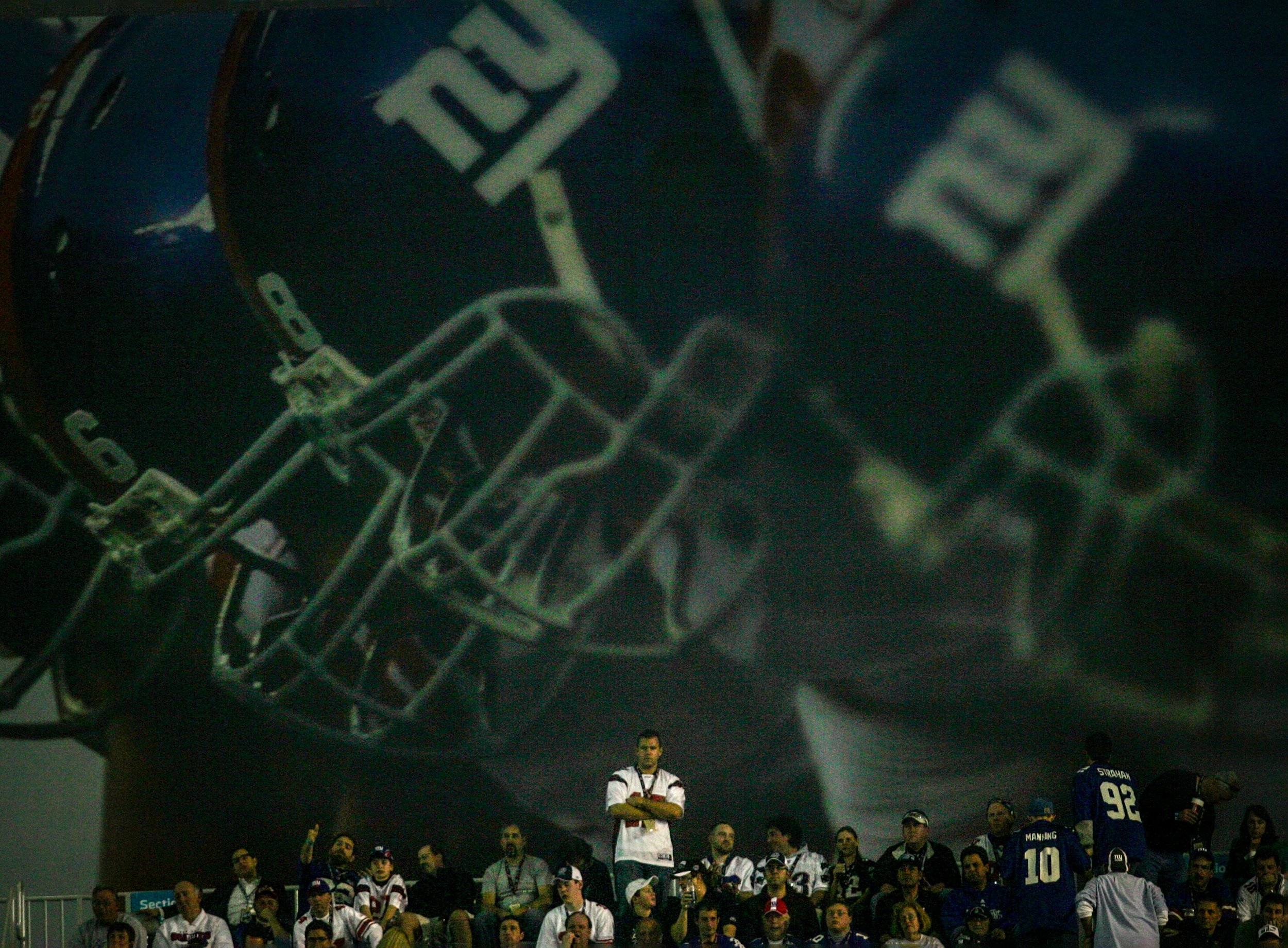 A nervous Patriot fan stands with his arms crossed in the back of the stands with a giant poster, showing Giantshelmets