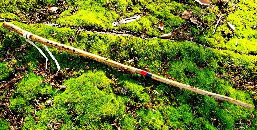 A wooden hiking stick flute lying on bright green moss and fallen leaves in a forest.