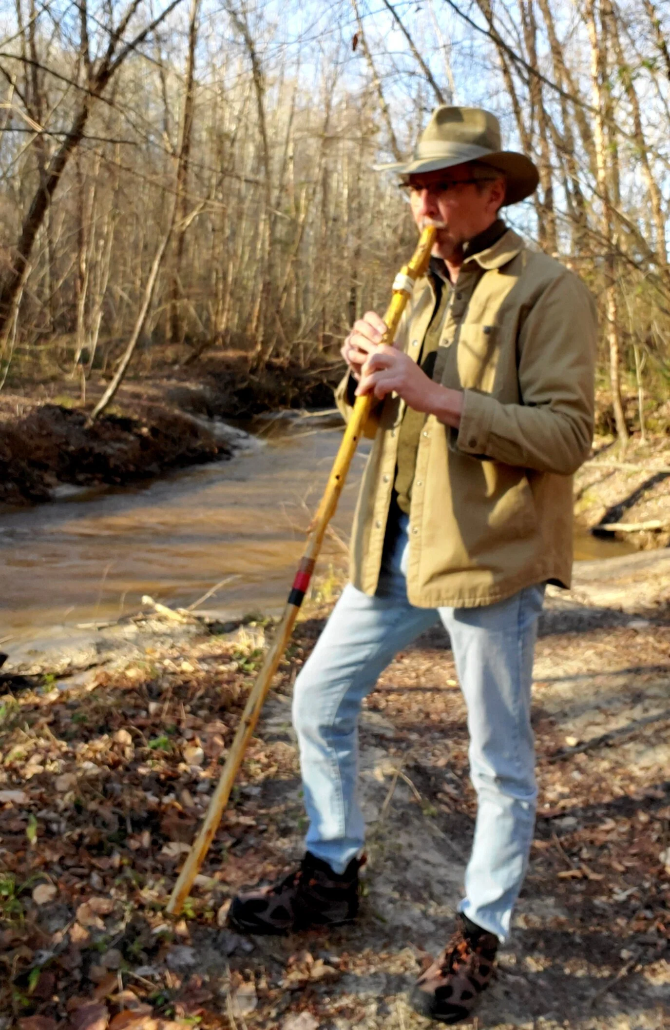 A man in outdoor clothing and a wide-brimmed hat standing in a wooded area near a stream, playing a walking stick flute.