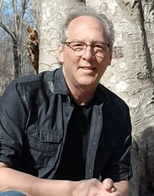 Man with glasses and gray hair smiling outdoors next to a tree.