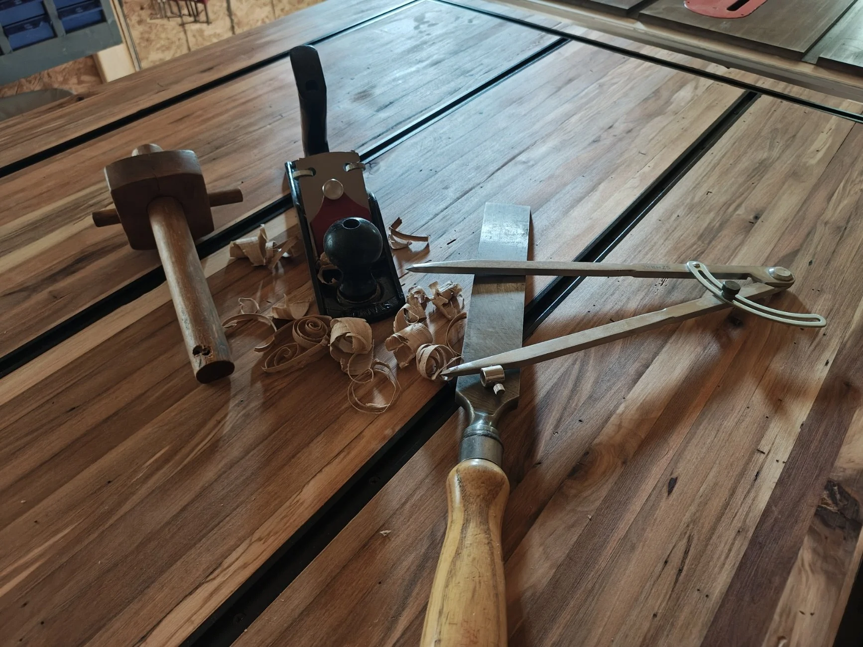 A woodworking workspace with a hand plane, a hacksaw, a hammer, a ruler, a square, wood shavings, and a wooden tabletop under construction.