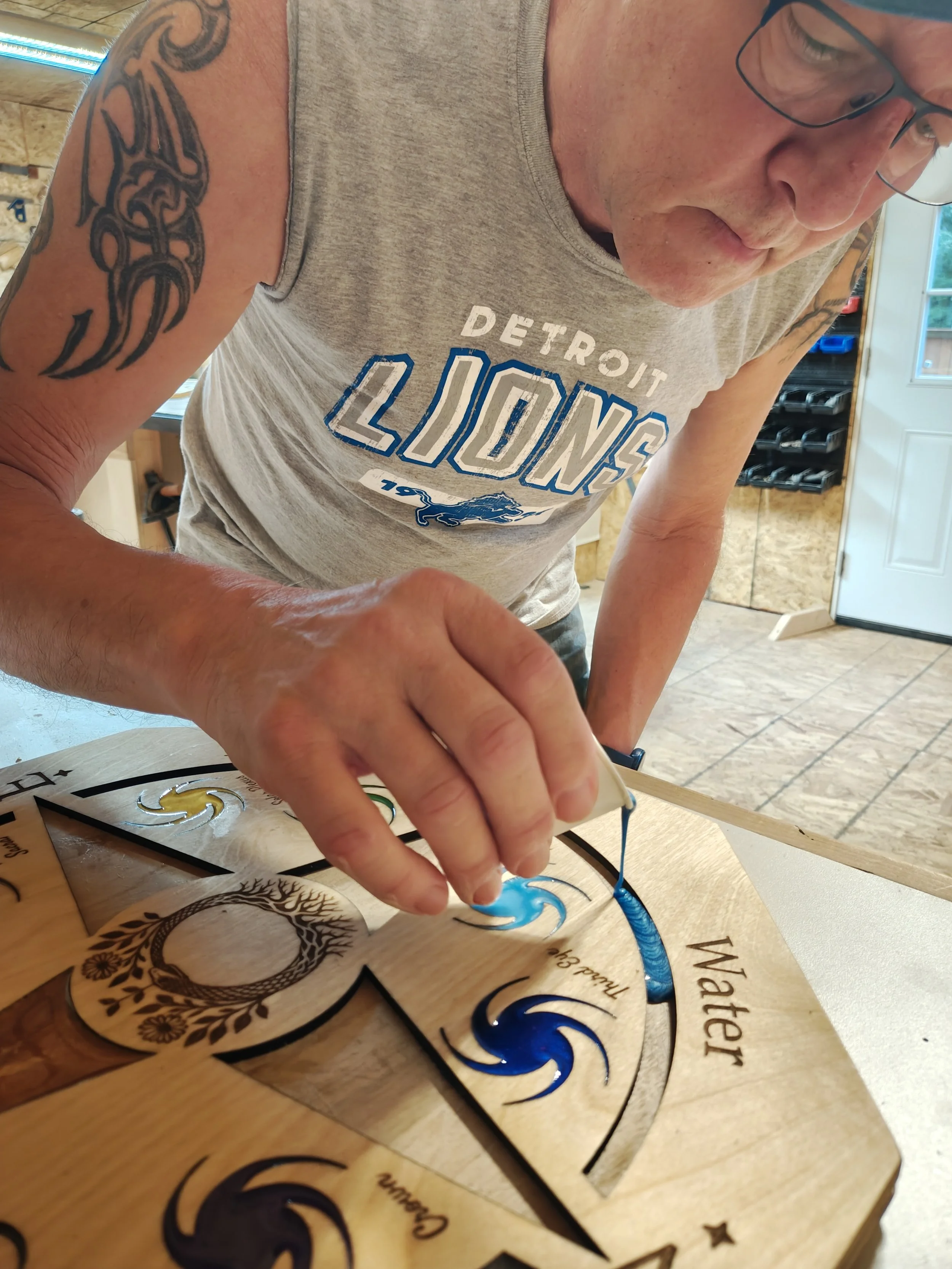 A man with tattoos on his arm, wearing a gray Detroit Lions tank top and glasses, is working on a woodcraft project with blue paint, in a workshop with wooden walls and a door.