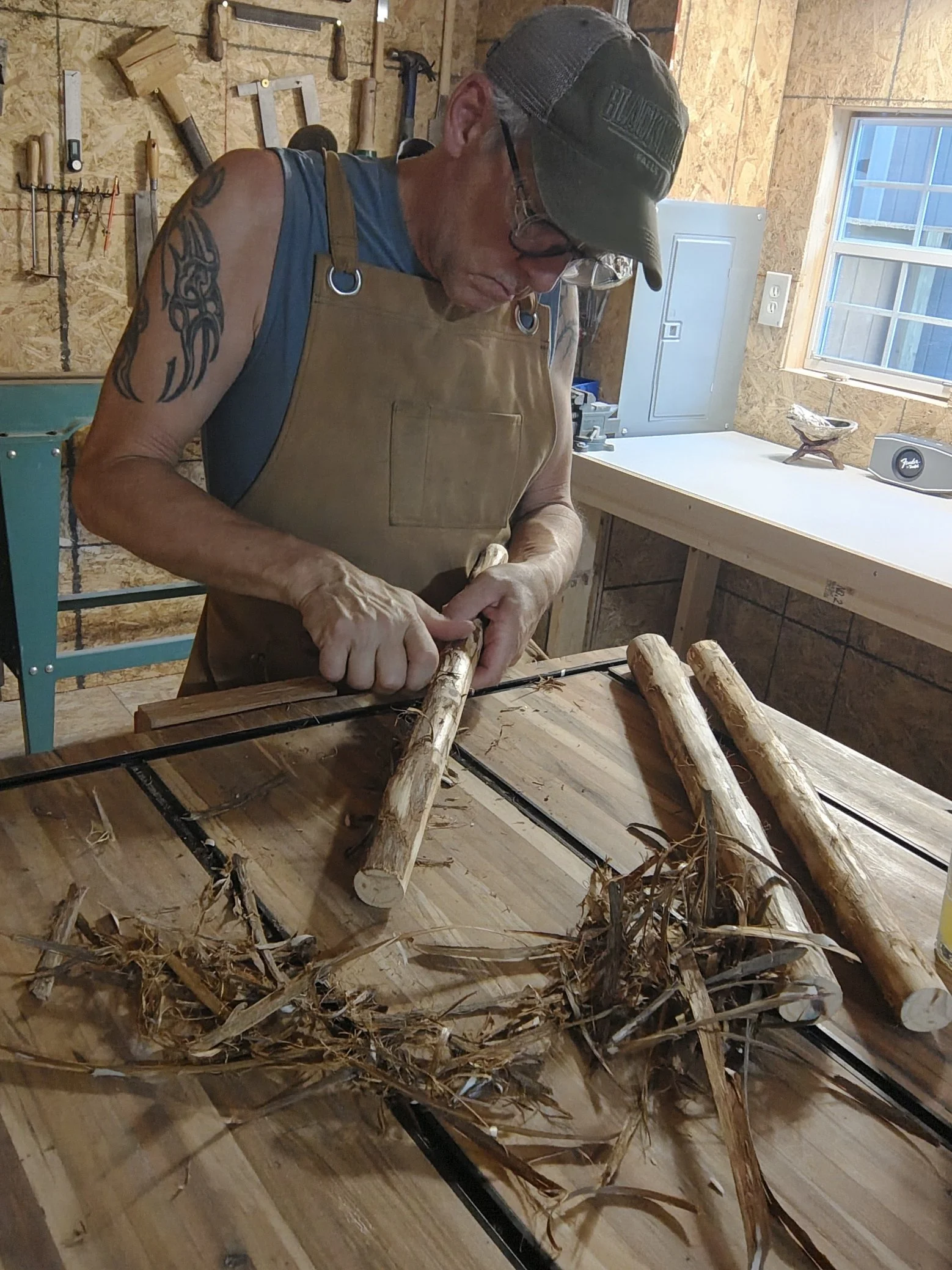 A person working in a woodworking shop, cutting and preparing wooden logs, with tools hanging on the wall and a window in the background.