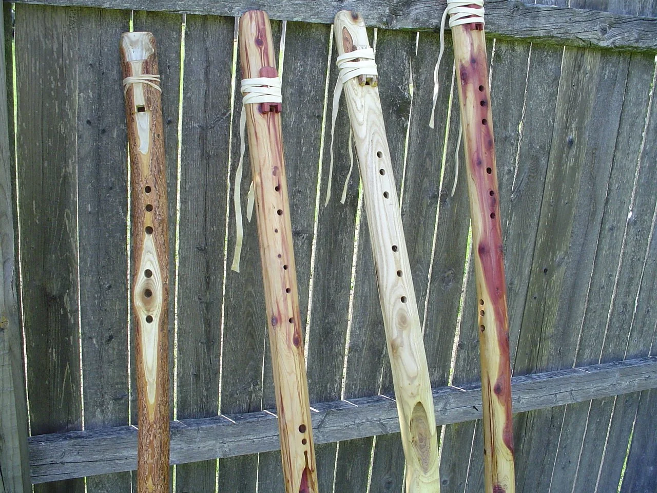 Four wooden native american style walking stick flutes leaning on a weathered wooden fence, each decorated with a leather strap at the top.