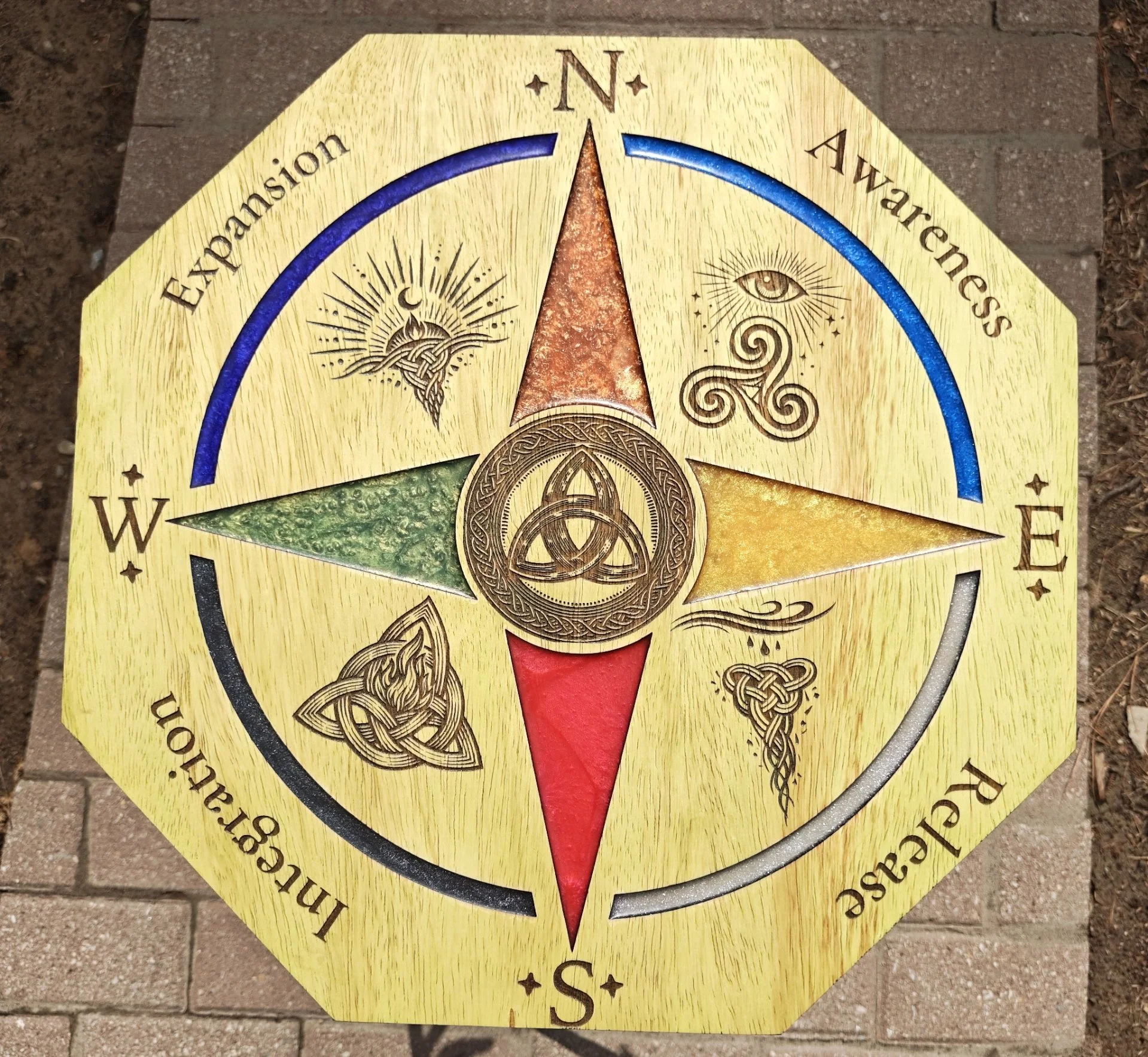A decorative directional sign with a Celtic knot design, a red downward arrow, and symbols for North, South, East, and West, on a brick surface.
