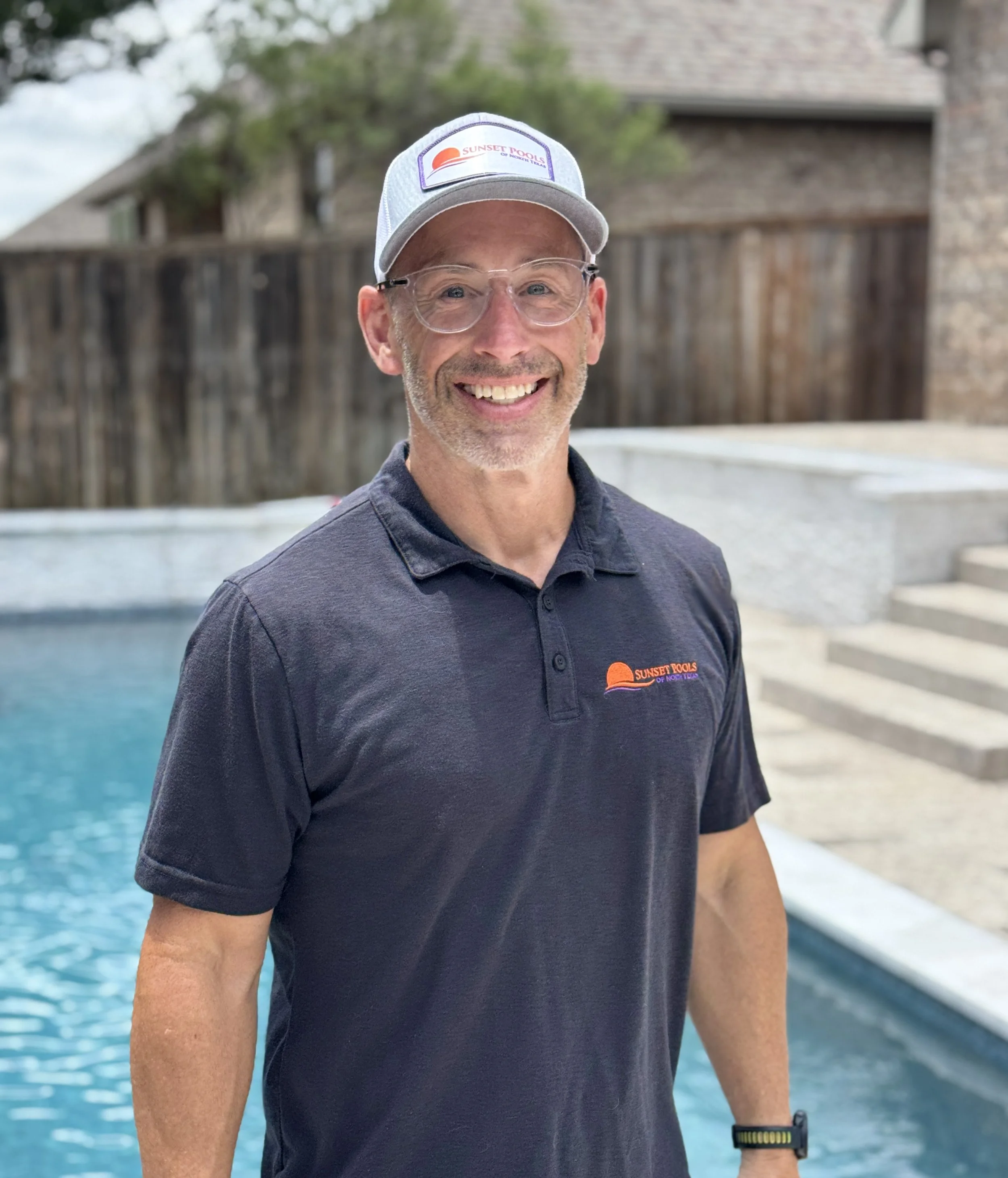 A smiling man wearing a gray baseball cap with a Sunset Pools logo, clear glasses, and a black polo shirt with a Sunset Pools logo, standing outdoors near a swimming pool with blue water and a wooden fence in the background.
