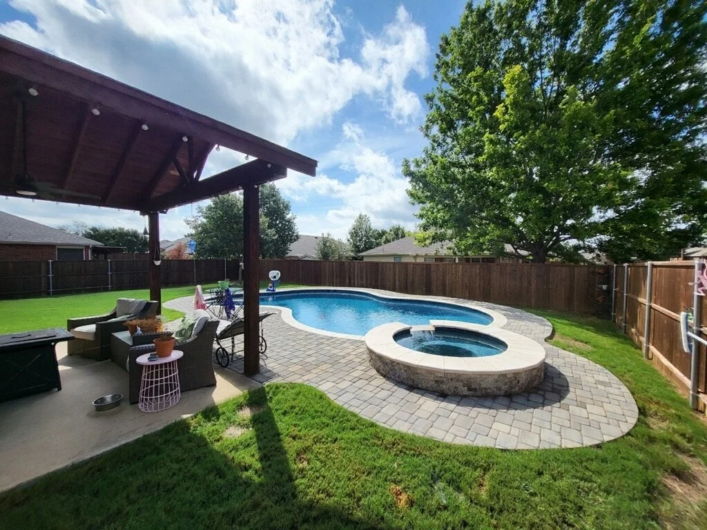 Backyard with an in-ground swimming pool, hot tub, patio furniture, and a wooden fence enclosing the yard with trees and houses in the background.