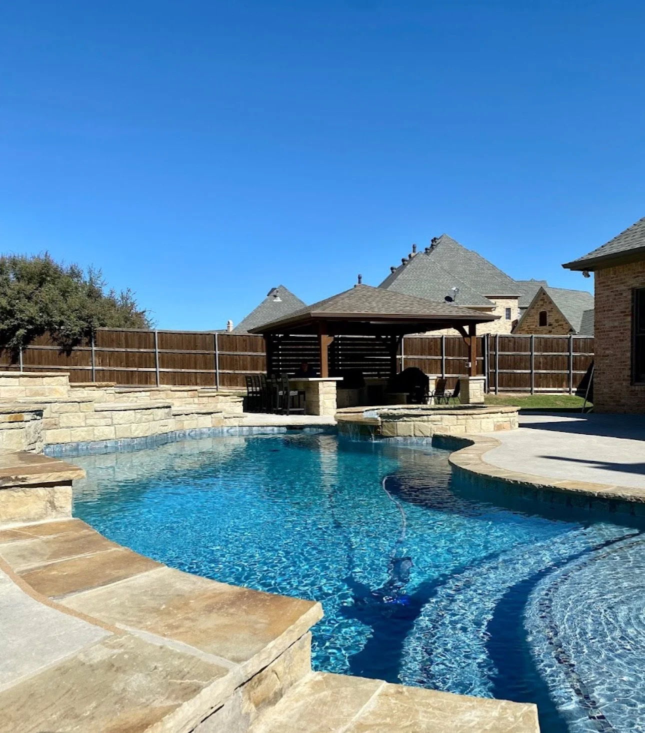 Backyard swimming pool with stone deck, surrounded by a wooden fence and a gazebo with outdoor seating, under a clear blue sky.