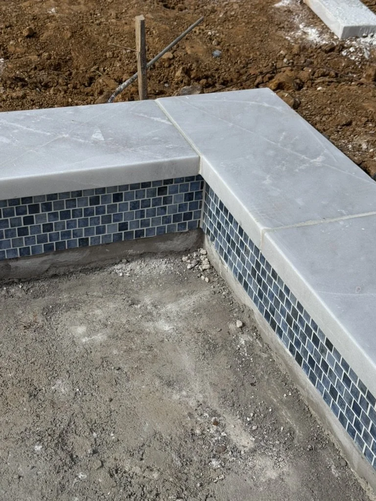 Corner of a swimming pool under construction with gray marble tiles on the edge and a black mosaic tile border. The surrounding area is dirt with some construction debris.