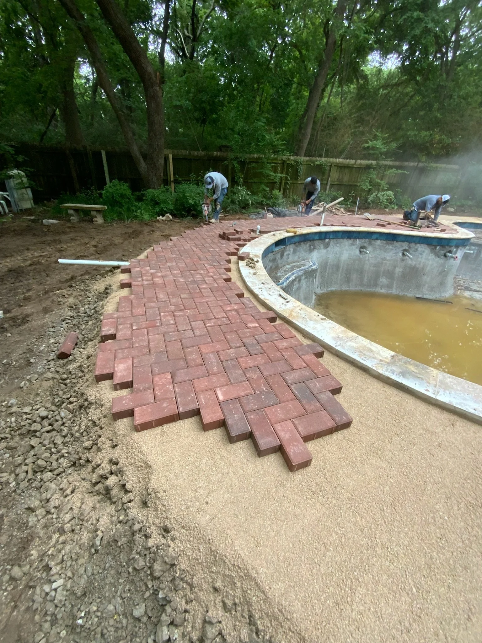 Workers installing a brick pathway around a pool in a backyard with trees and a wooden fence.