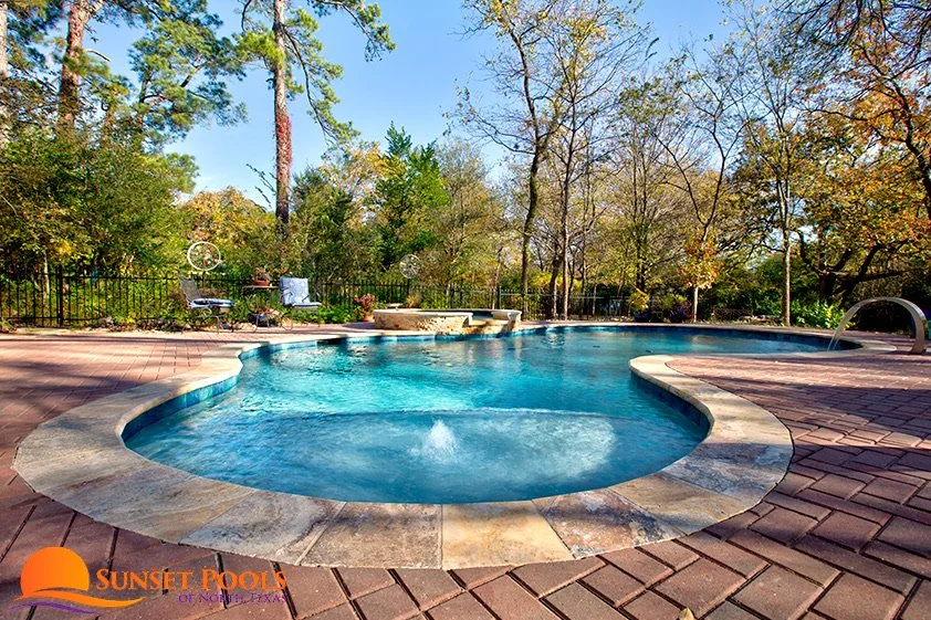 A backyard with a kidney-shaped swimming pool surrounded by a brick patio and trees with autumn foliage, featuring a hot tub at one end and a fountain at the far end.