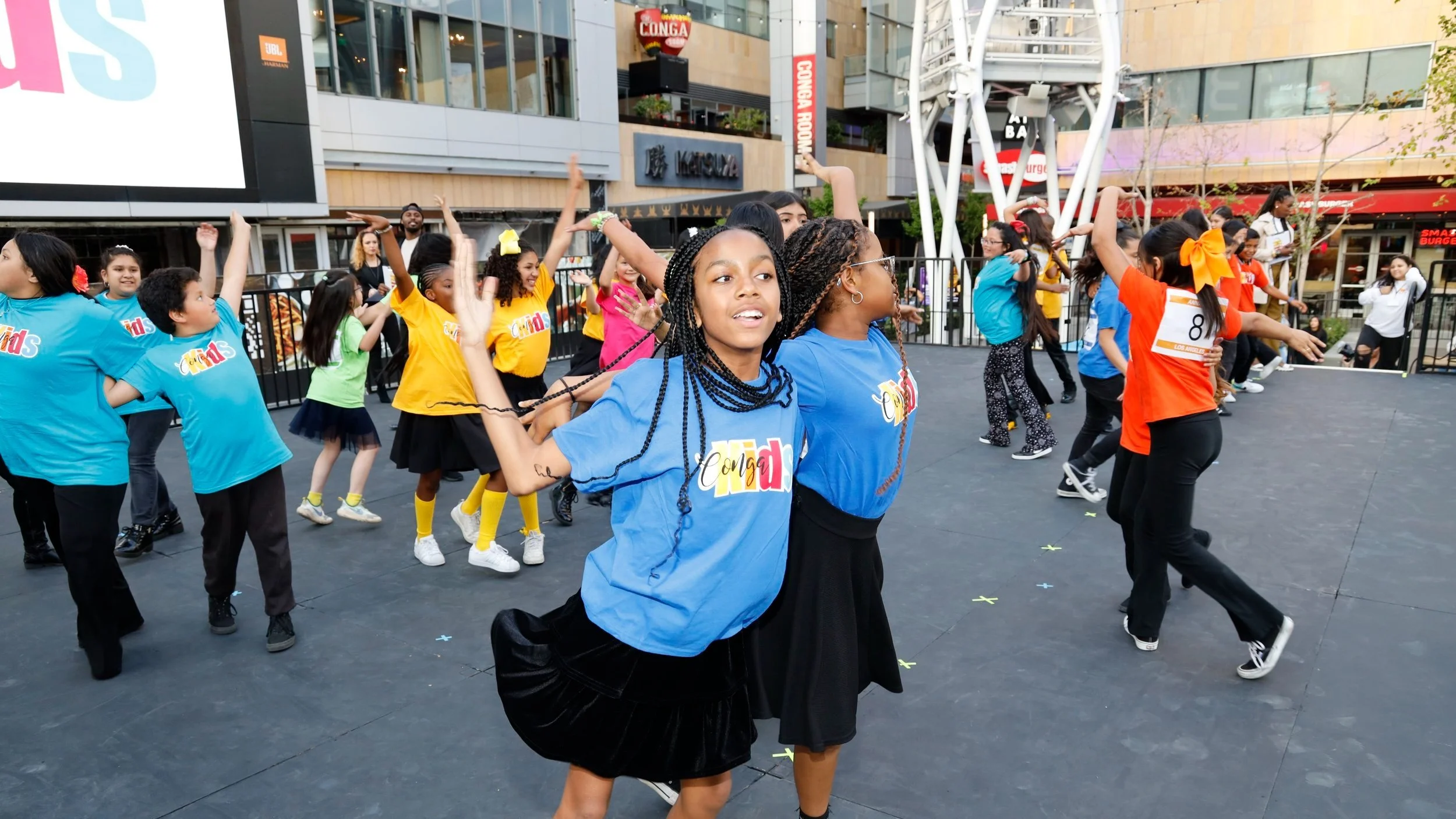 Group of children participating in a dance or exercise activity outdoors in an urban setting, wearing colorful shirts with 'Conça' printed on them, with some children in blue, yellow, and orange shirts, and a Ferris wheel in the background.