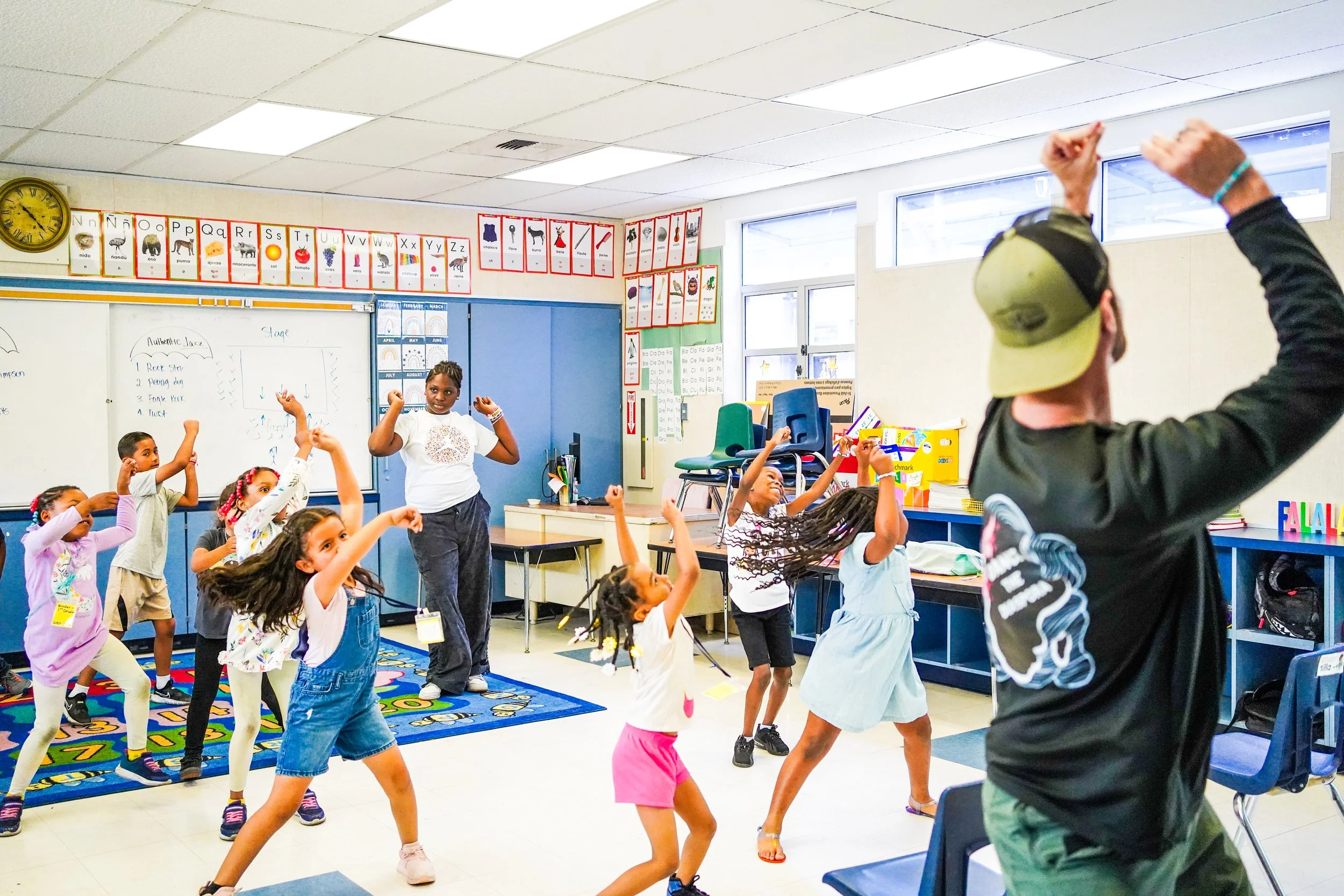 A diverse group of young children and a teacher are participating in a dance activity in a classroom. The children are moving with raised arms and appear to be enjoying themselves.