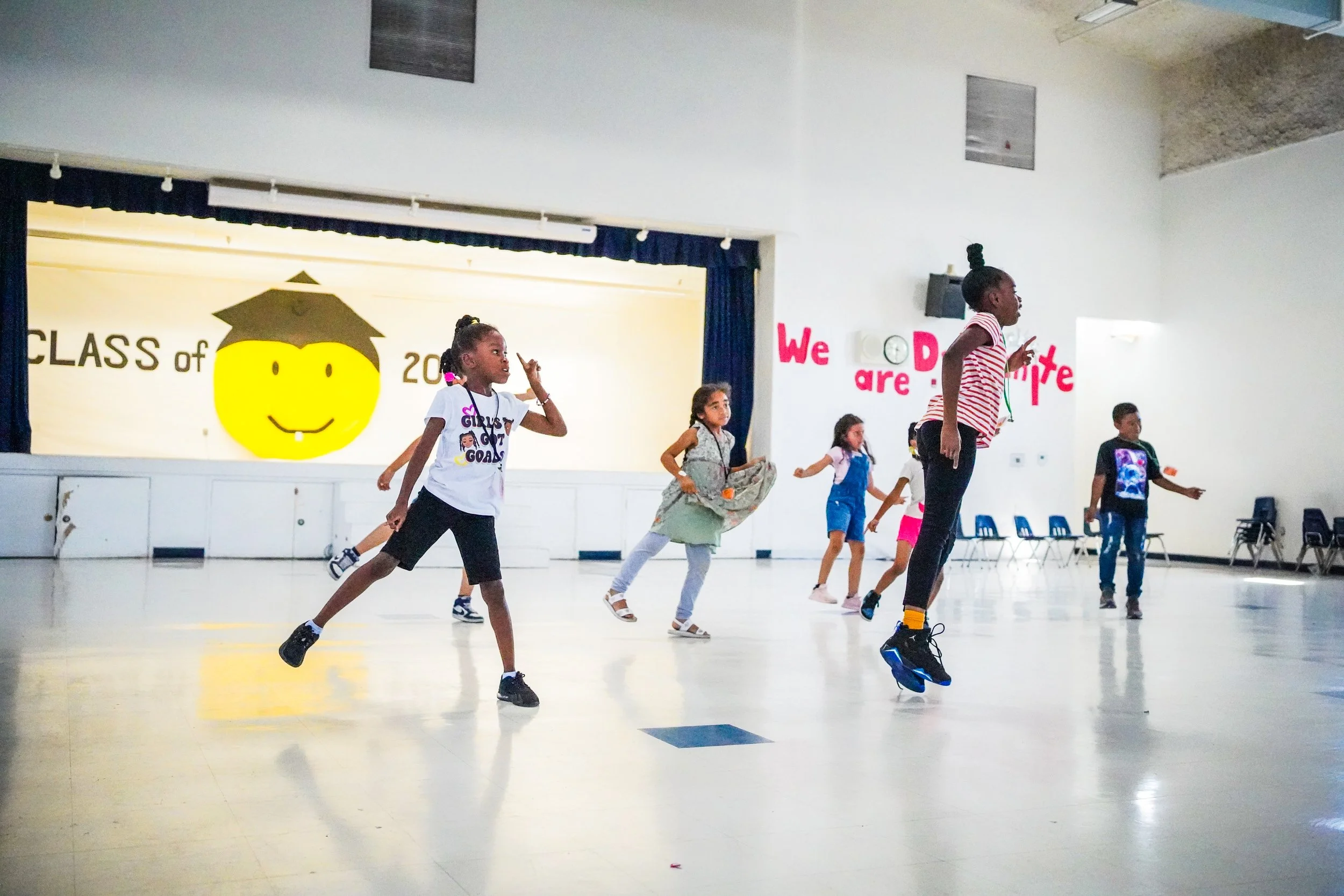 Children dancing and jumping in a school auditorium with a decorated stage and a sign that reads 'We are D' at the back.