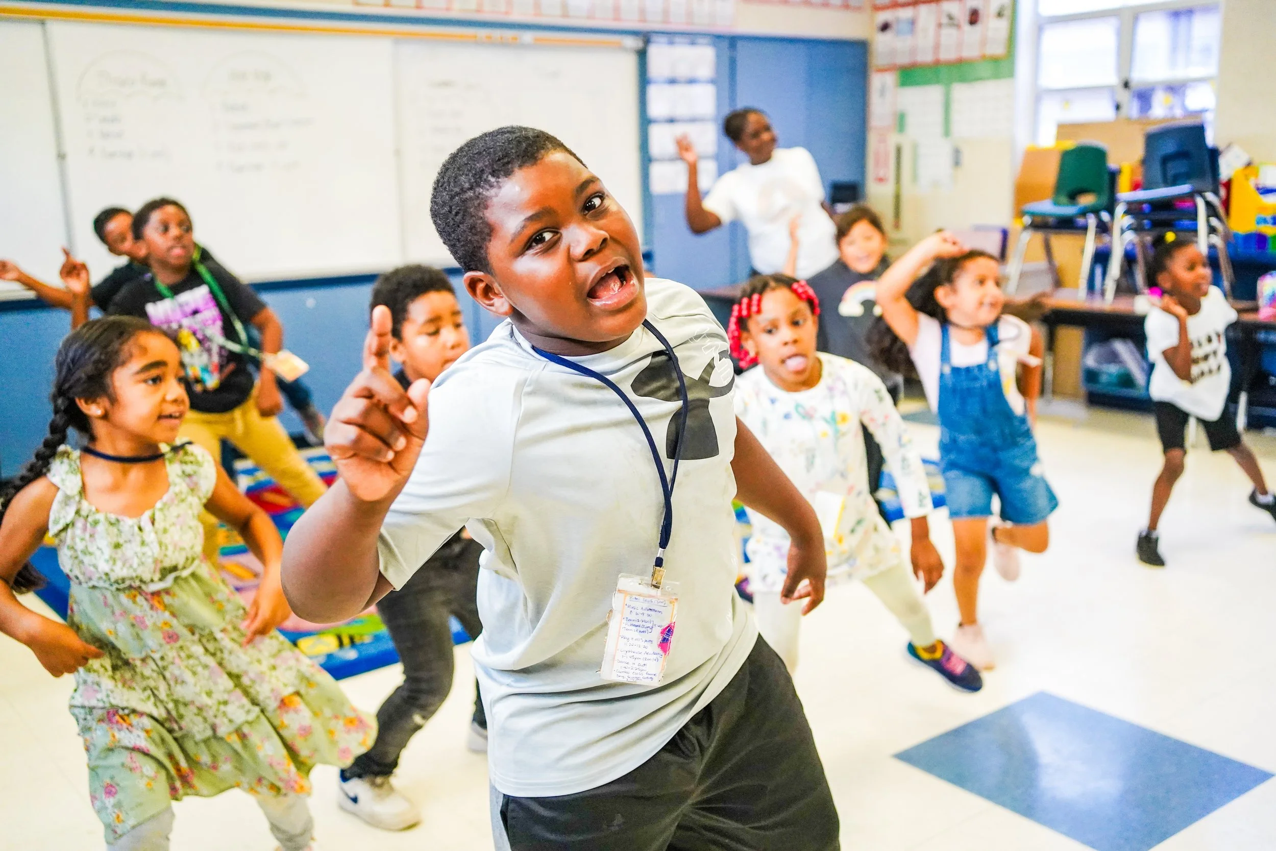 Young boy leading a dance activity in a classroom with young children and a teacher in the background.