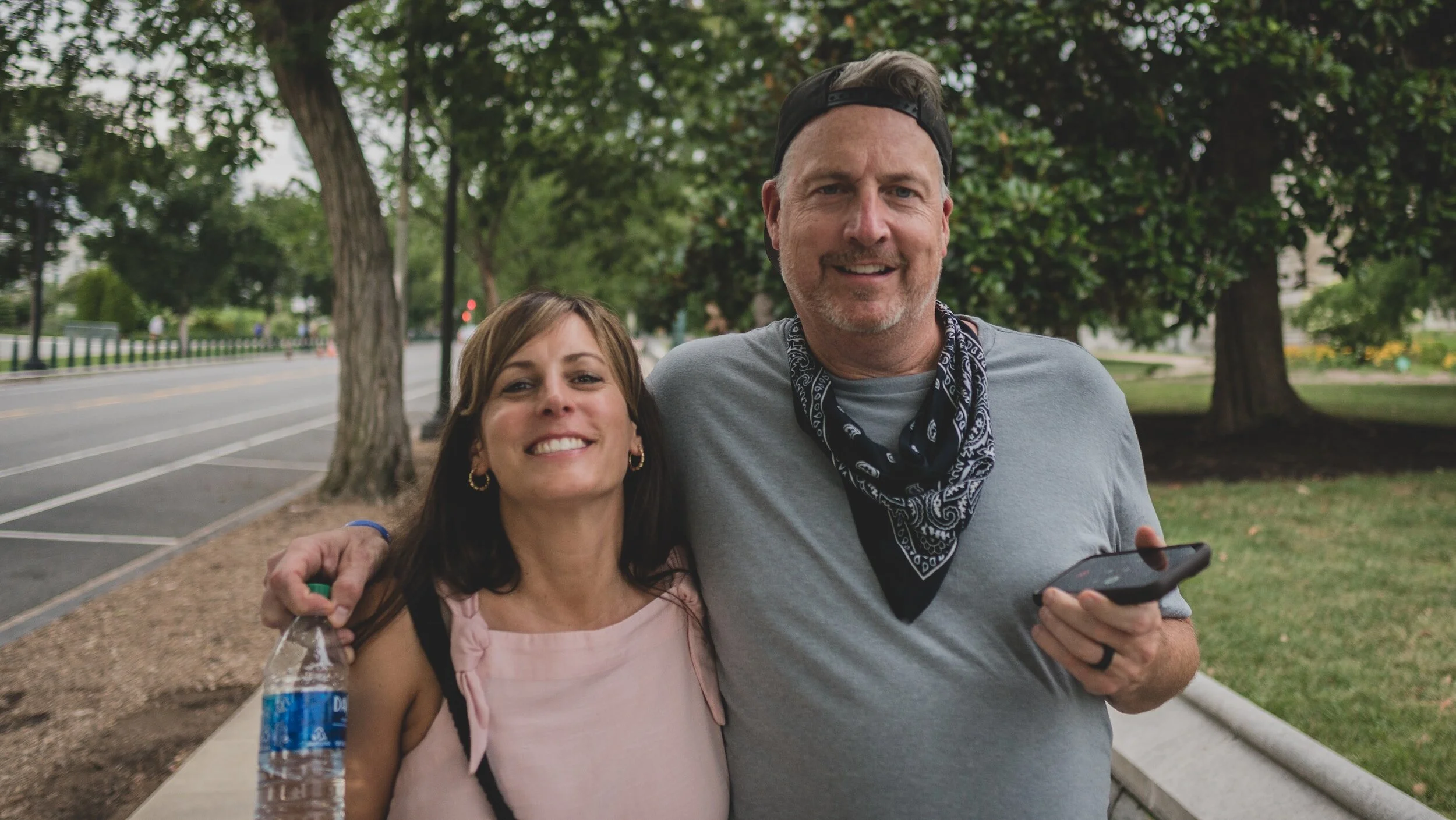 Couple walking, gentleman has arm over ladies shoulder he is holding a phone in one hand and a water bottle in the other
