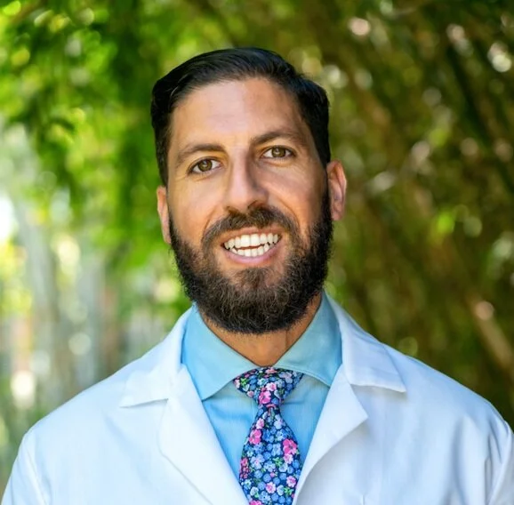 A man with dark hair, a beard, and a friendly smile, wearing a white coat over a light blue shirt and a floral tie, standing outdoors with blurred green foliage in the background.