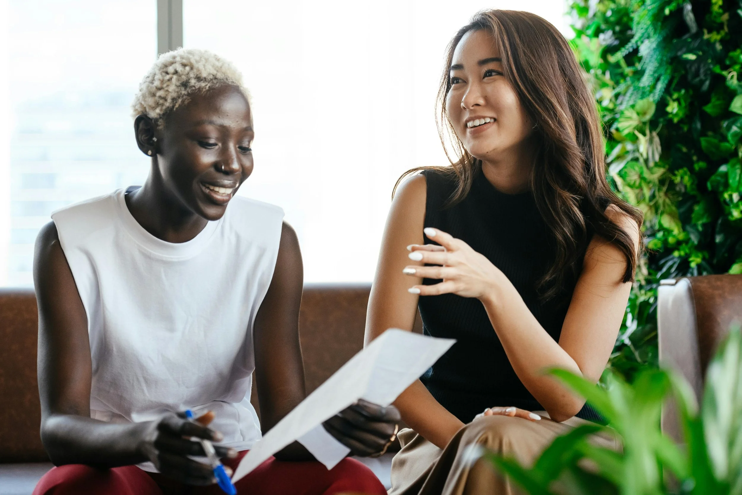 Two women sitting and talking in a bright room. One woman has short bleached hair and is holding a paper and pen, smiling. The other woman has long brown hair and is gesturing with her hand, smiling. There is greenery in the background.