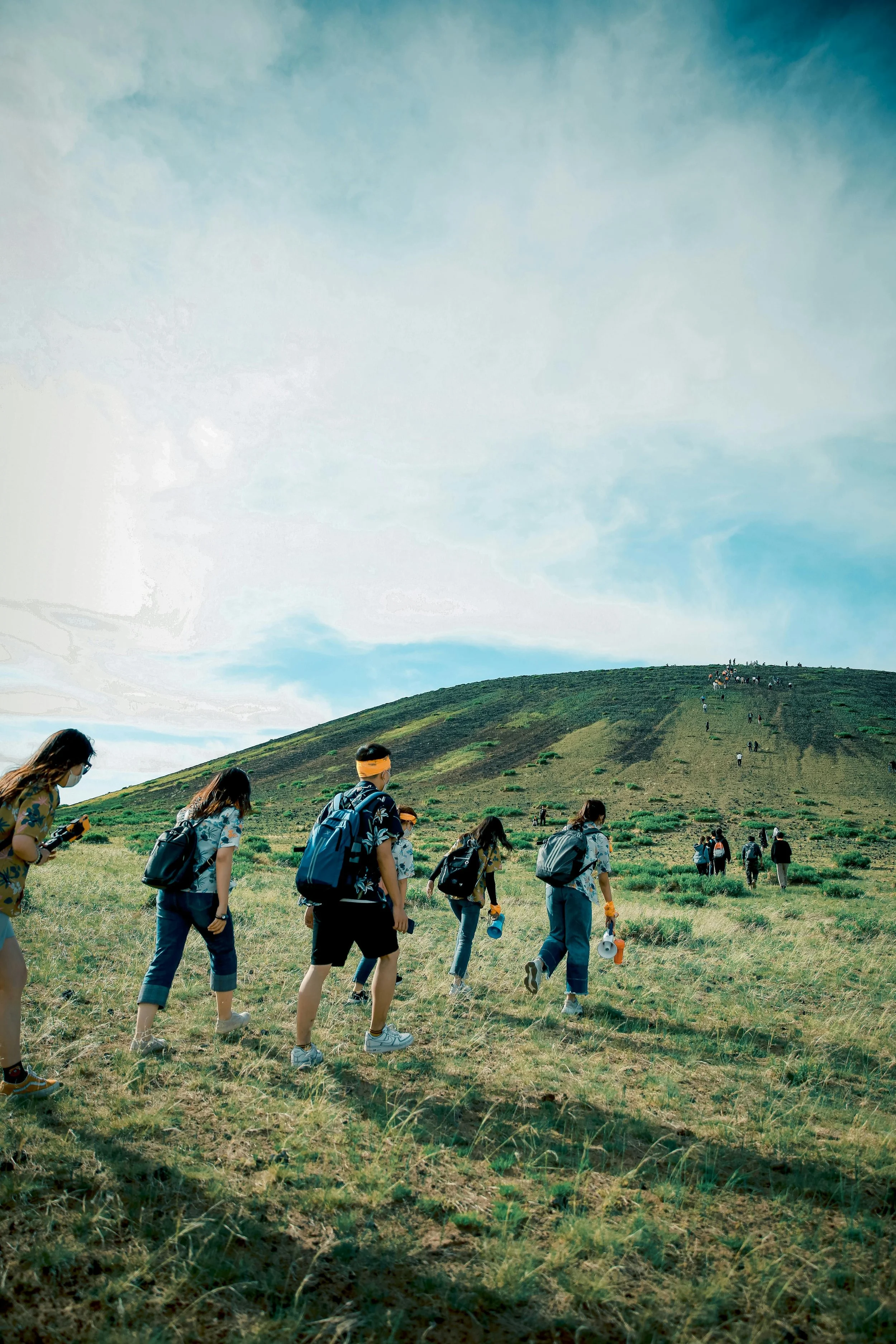 Group of people hiking up a grassy hill under a partly cloudy sky, some carrying backpacks and water bottles.