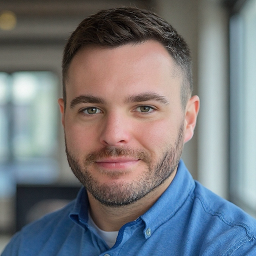 Close-up portrait of a young man with short dark hair and a beard, wearing a blue collared shirt, smiling mildly, in an indoor setting with blurred background.