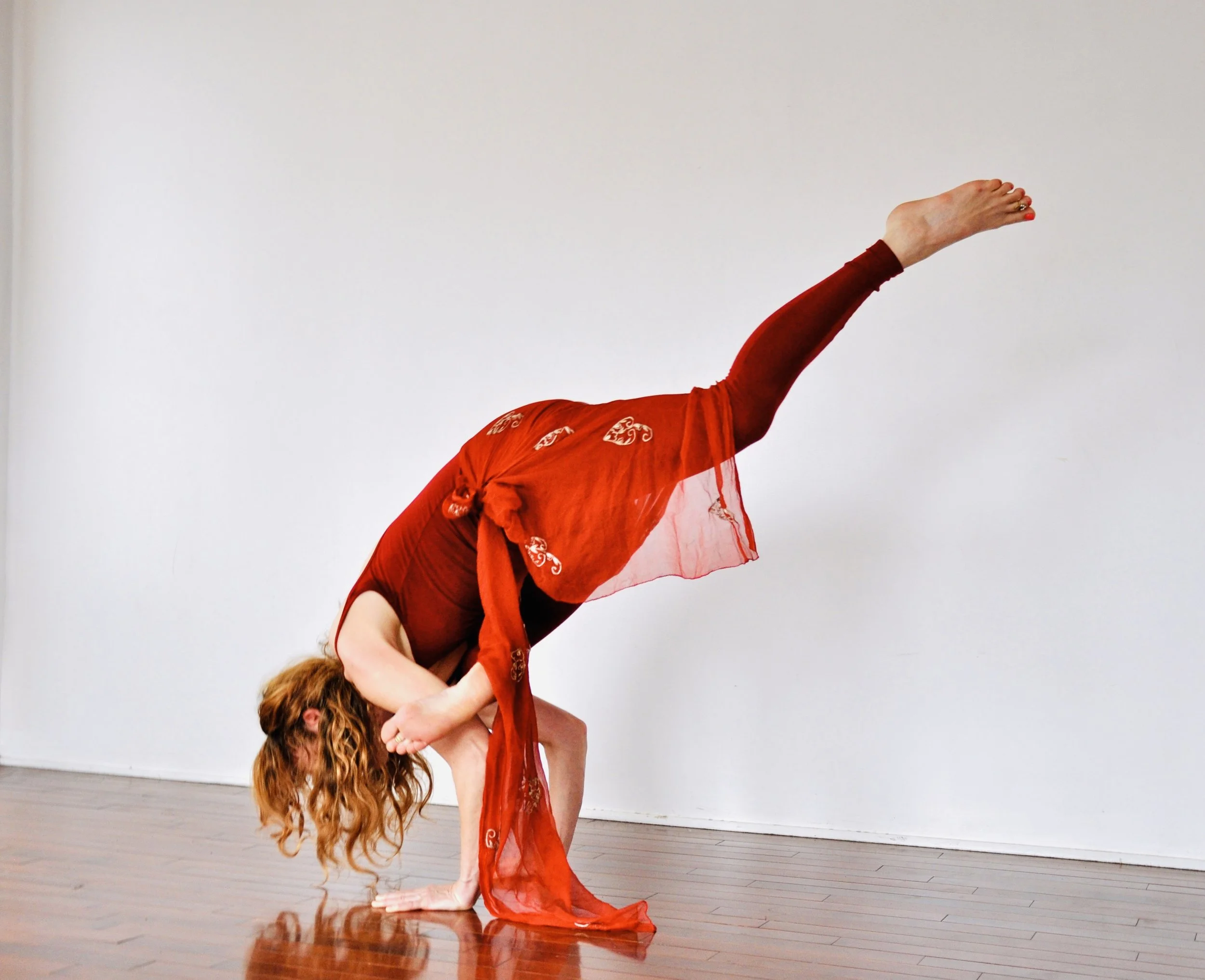 Woman in red dress performing a yoga pose on a wooden floor with a white wall background.