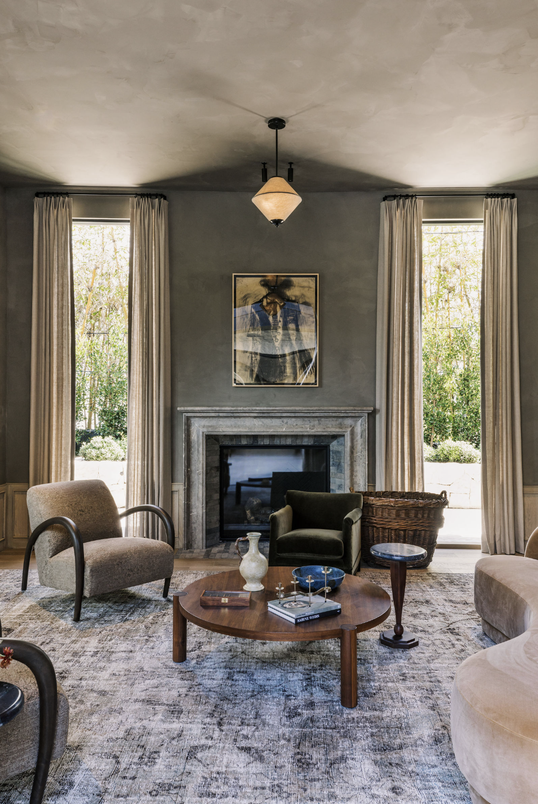 Living room with gray walls, two tall windows with beige curtains, a fireplace, modern art above the fireplace, a round wooden coffee table with decorative items, upholstered armchairs, a patterned rug, and greenery visible outside.