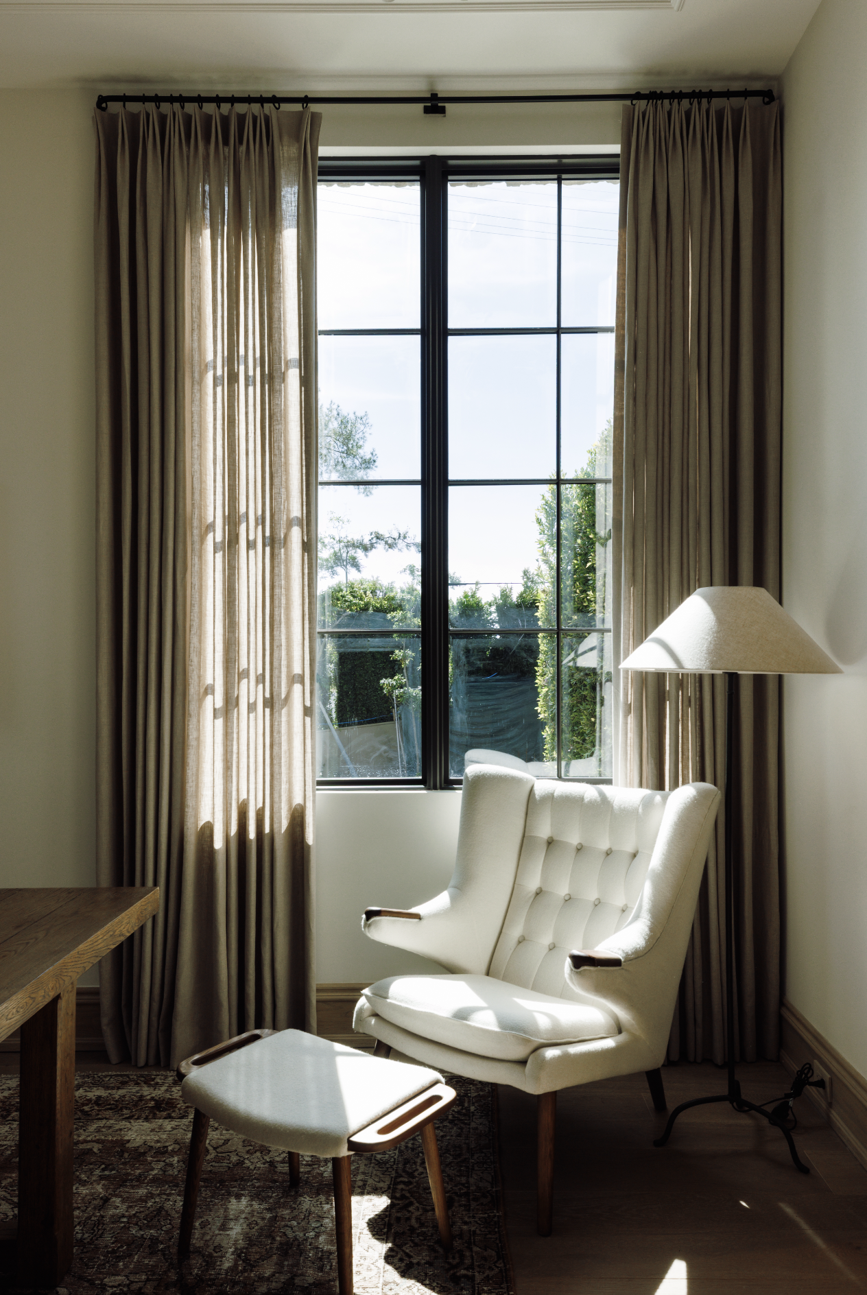 A cozy room with beige curtains, a large black-framed window with sunlight streaming in, a cream-colored armchair with a matching footstool, a table, a floor lamp, and a patterned rug.