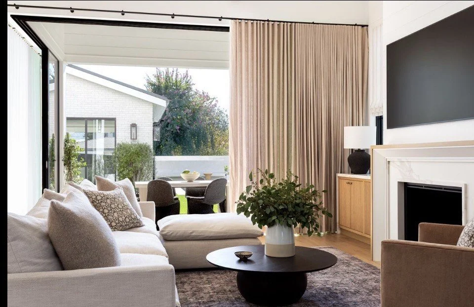 Bright living room with white sofa, patterned pillows, black coffee table with plant, beige curtains, fireplace, gray accent wall, and an outdoor dining area with black chairs and a white table visible through sliding glass door.